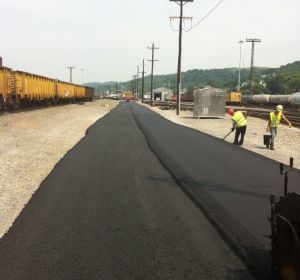 Workers paving a black road beside parked yellow freight train cars in an industrial yard