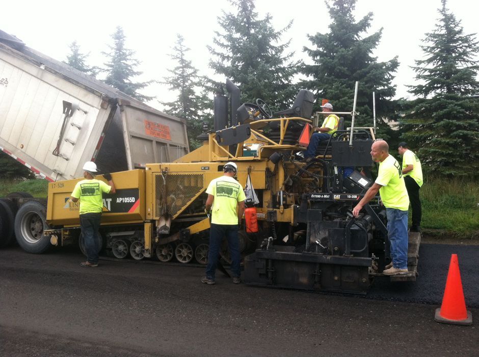 Road crew with yellow pavers working beside a white dump truck on a wet street