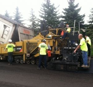 Workers operate a yellow road machine beside a dump truck on a paved road under cloudy skies.