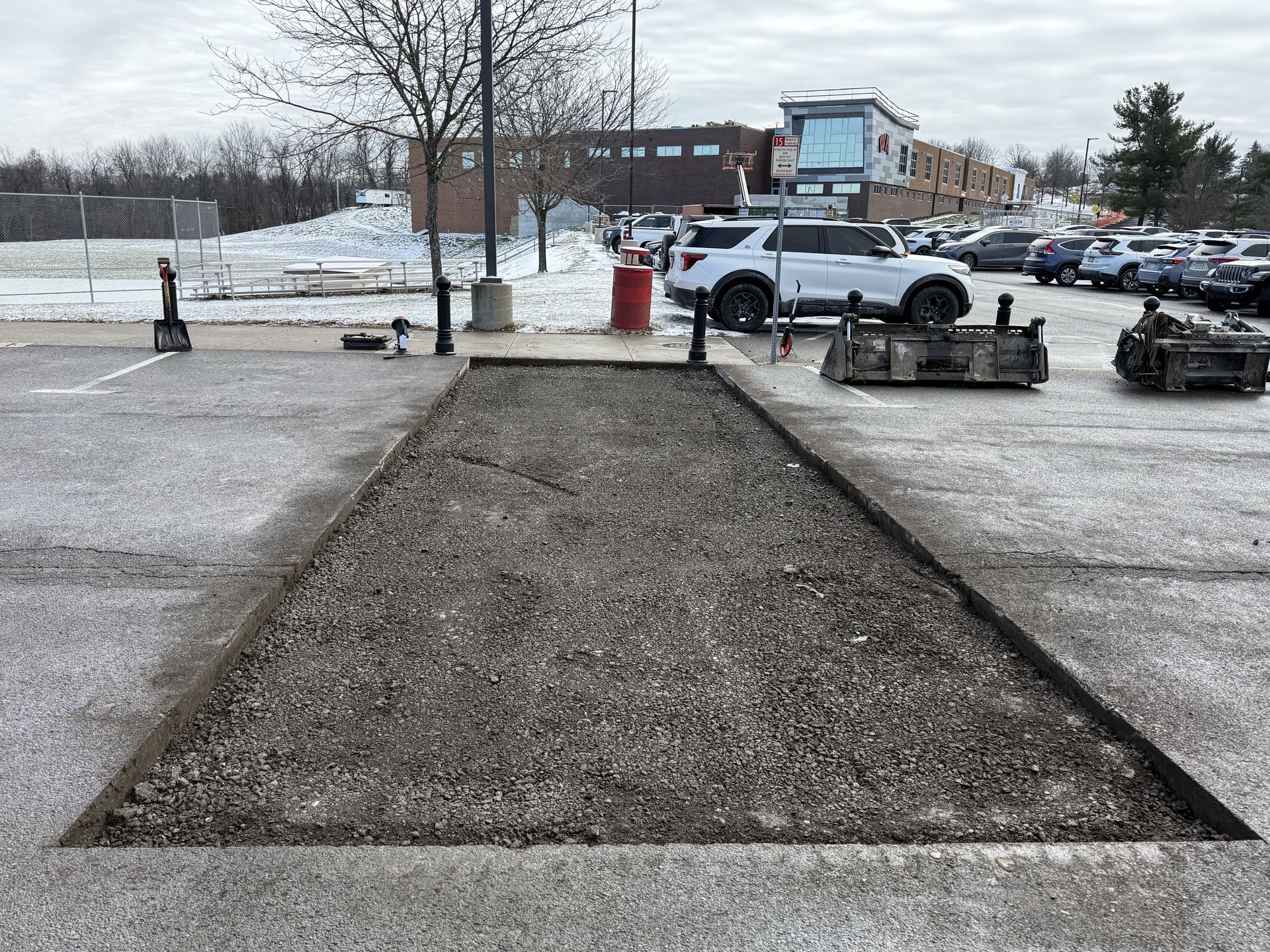 Snow-covered parking lot with a gravel path leading toward a building, with parked vehicles and trees in the background