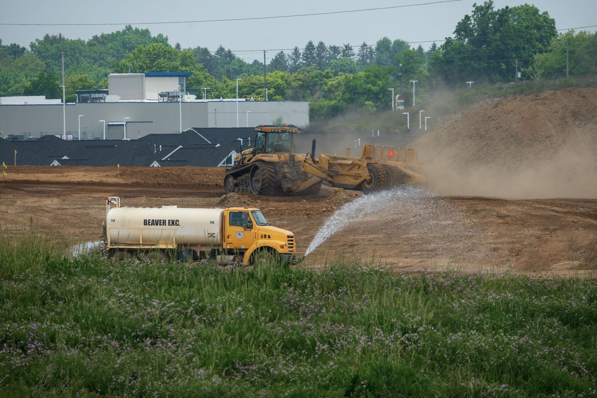 Construction site with a yellow water truck spraying dirt beside heavy equipment and dust clouds