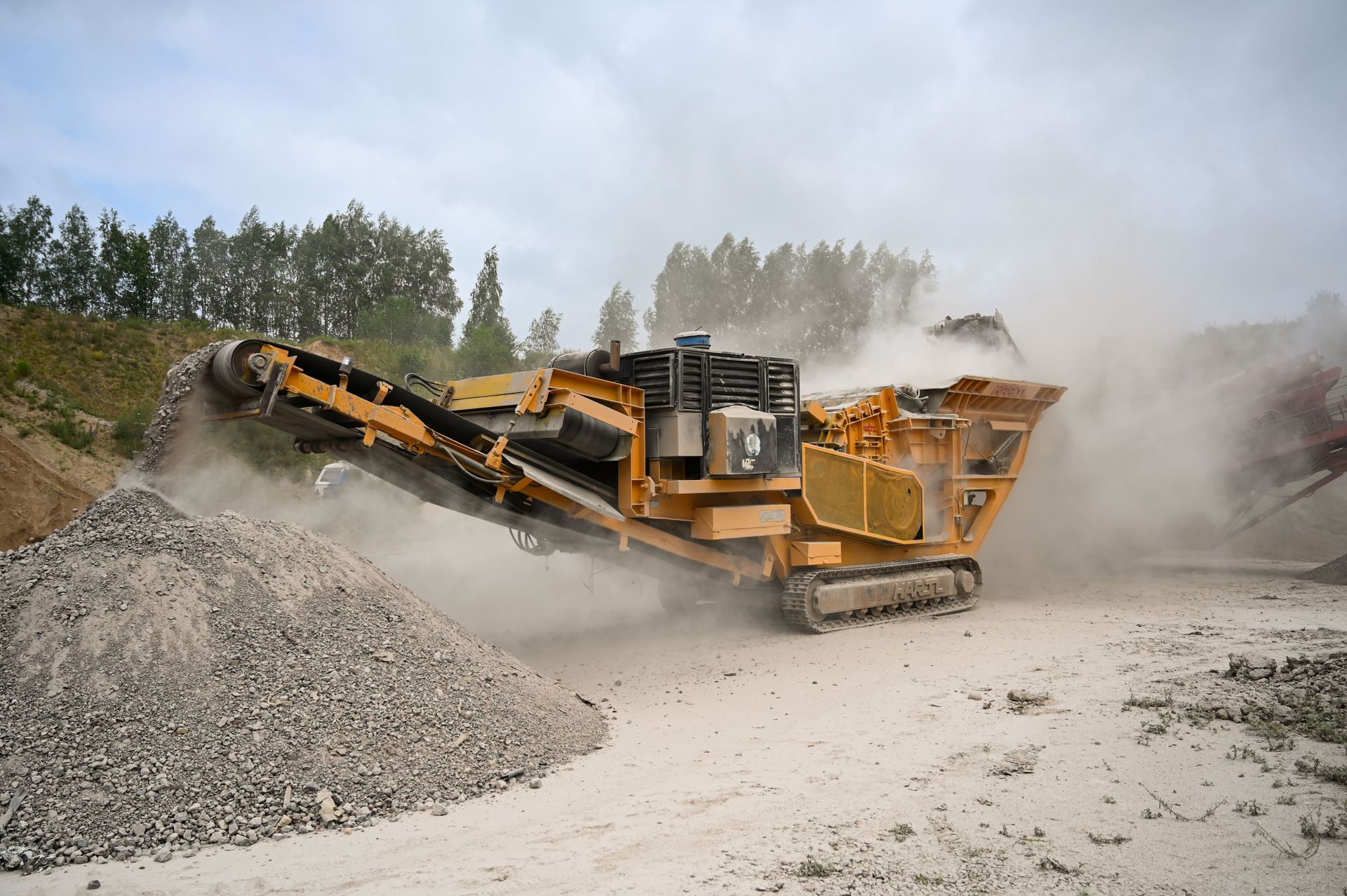 Large yellow crusher machine on a dusty gravel site with piles of rock and trees in the background