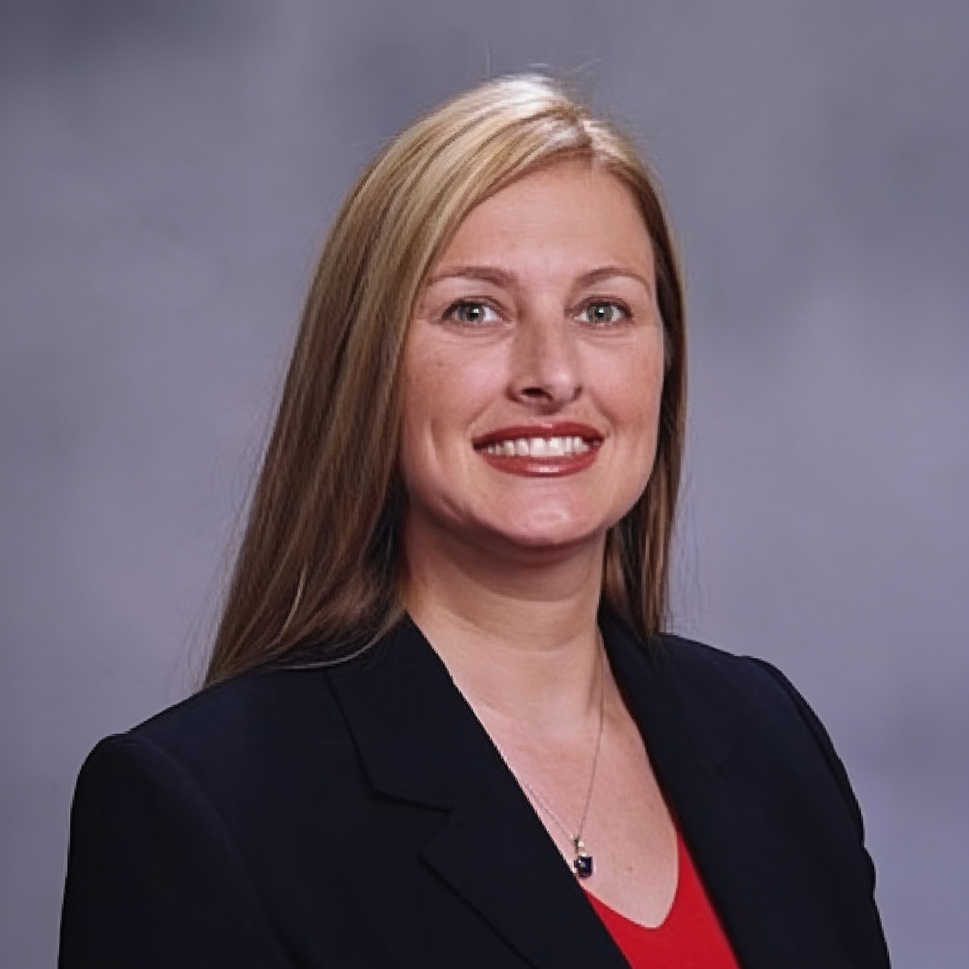 Professional headshot of a smiling woman in a black blazer and red top against a gray background