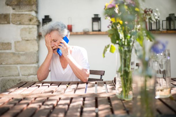 Woman seated at an outdoor style table, upset and on the phone next to a vase of flowers.