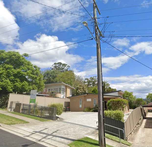Exterior view of a building with a paved driveway, trees, and a utility pole against a blue sky.