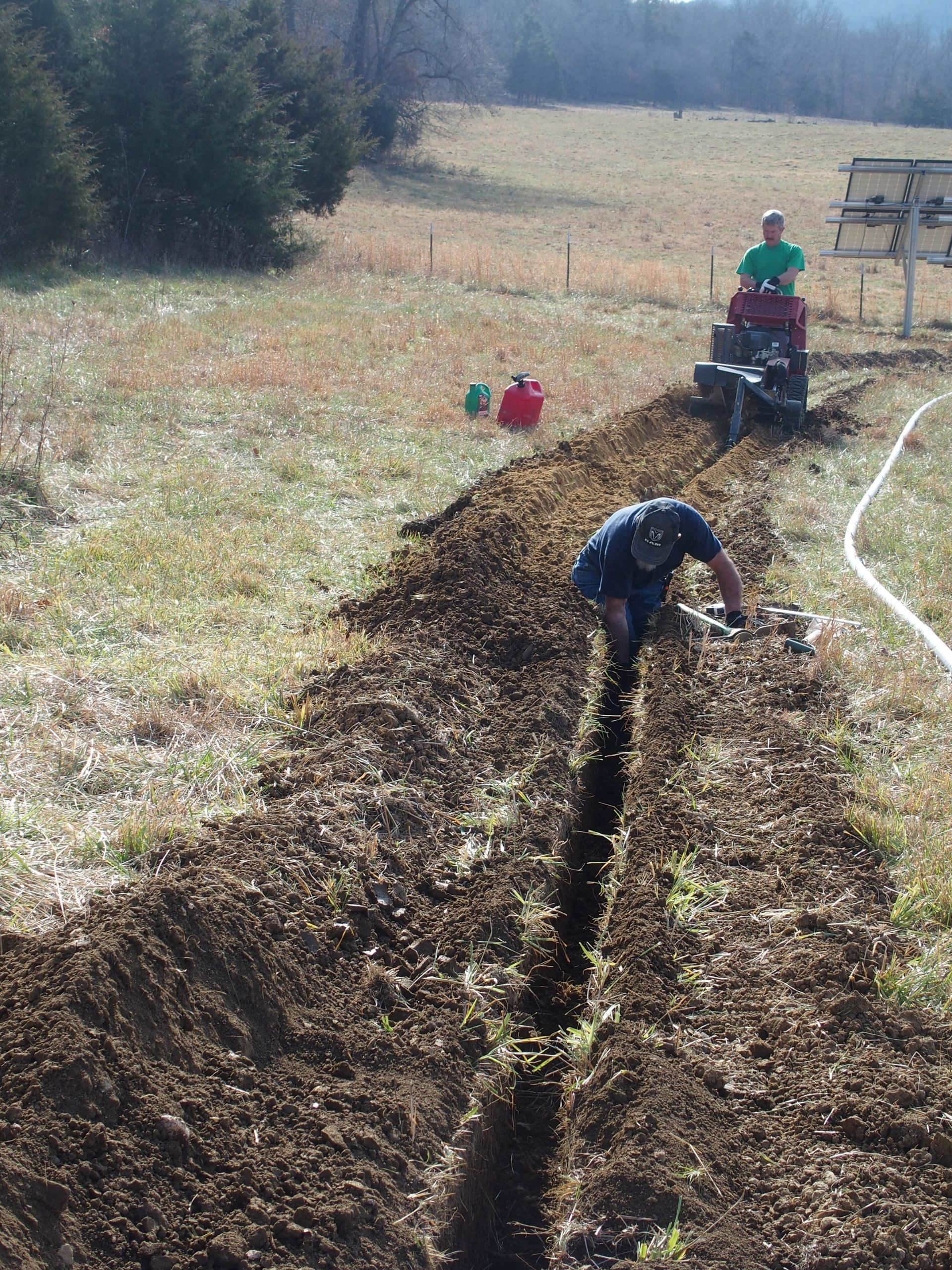 A man is digging a trench in a field with a tractor in the background.