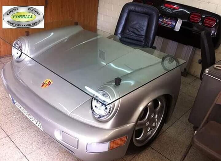 La parte delantera de un Porsche plateado, reconvertida en escritorio con superficie de cristal, se encuentra en una habitación con una silla negra.