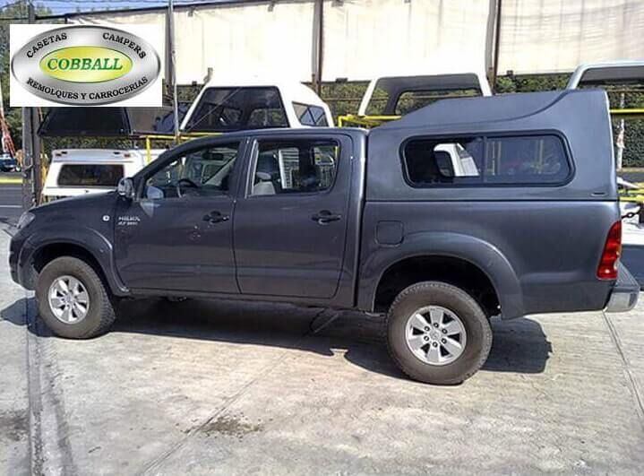 Una camioneta Toyota Hilux de color gris oscuro con una capota a juego, estacionada en un aparcamiento al aire libre con un letrero de Cobball en una esquina.