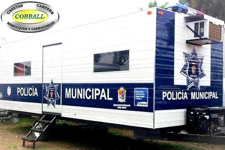 Unidad móvil de la Policía Municipal, de color blanco y azul, montada sobre un remolque con logotipo, puerta, ventanas y unidad de aire acondicionado.
