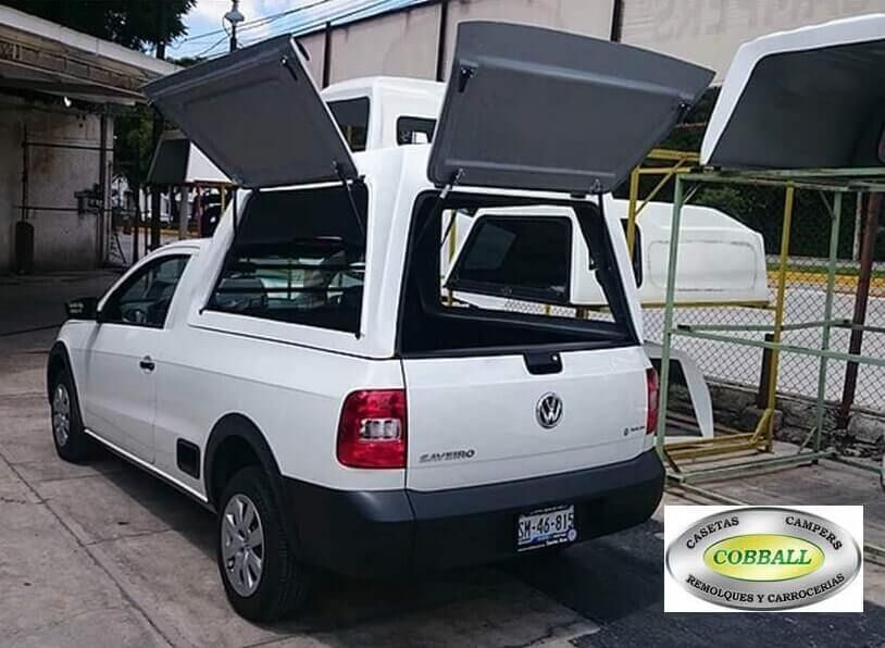 Una camioneta Volkswagen Saveiro blanca, con la cubierta de carga trasera abierta, estacionada en un área de taller al aire libre.
