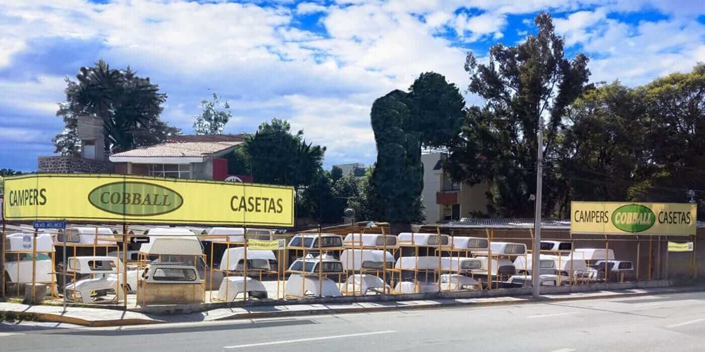 Un terreno cercado al aire libre donde se exhiben muchas cubiertas blancas para camionetas camper bajo letreros amarillos que dicen 