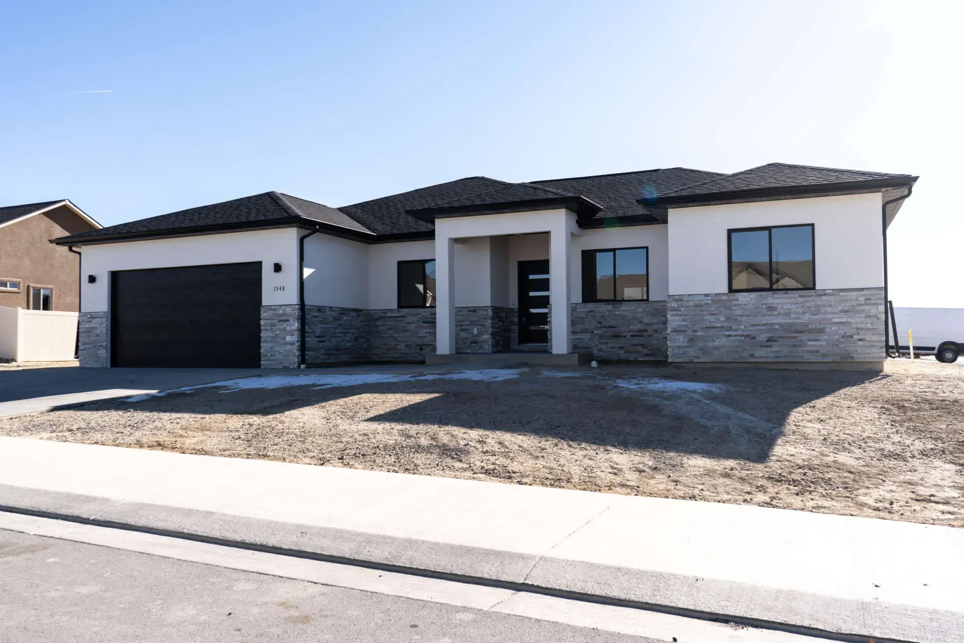 A single-story modern house with a light stucco finish, stone veneer base, black roof, and an attached double garage.