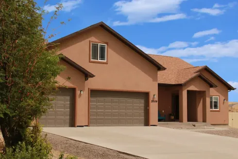 A tan, single-story stucco house with a three-car garage and brown tiled roof under a blue sky.