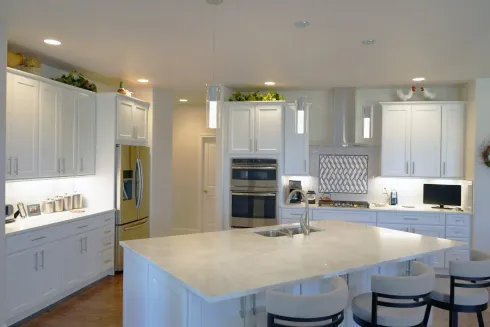 A modern white kitchen featuring a large island with three chairs, stainless appliances, and pendant lighting.