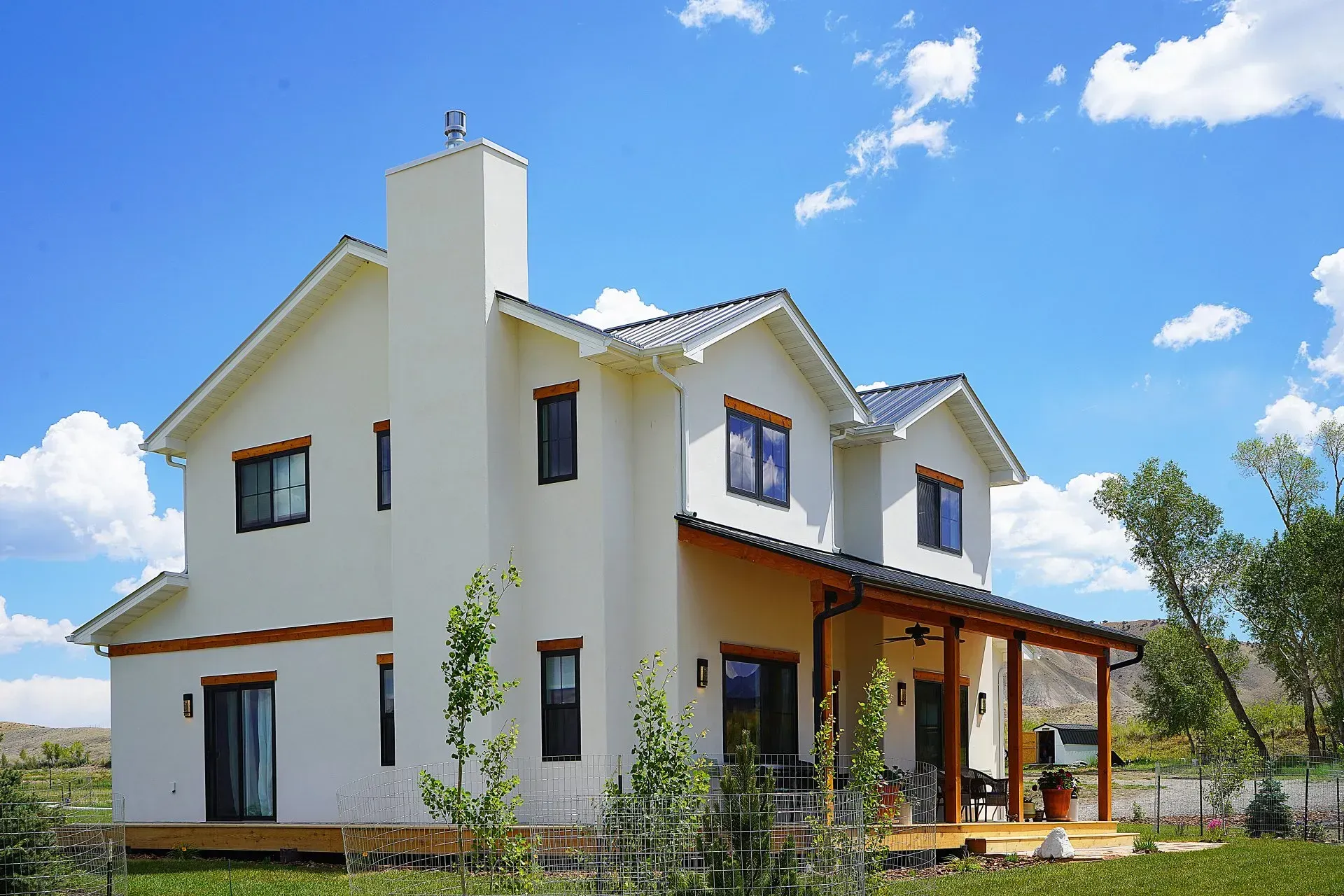 A two-story, cream-colored stucco home with a dark roof and a wooden front porch, set under a blue sky with clouds.