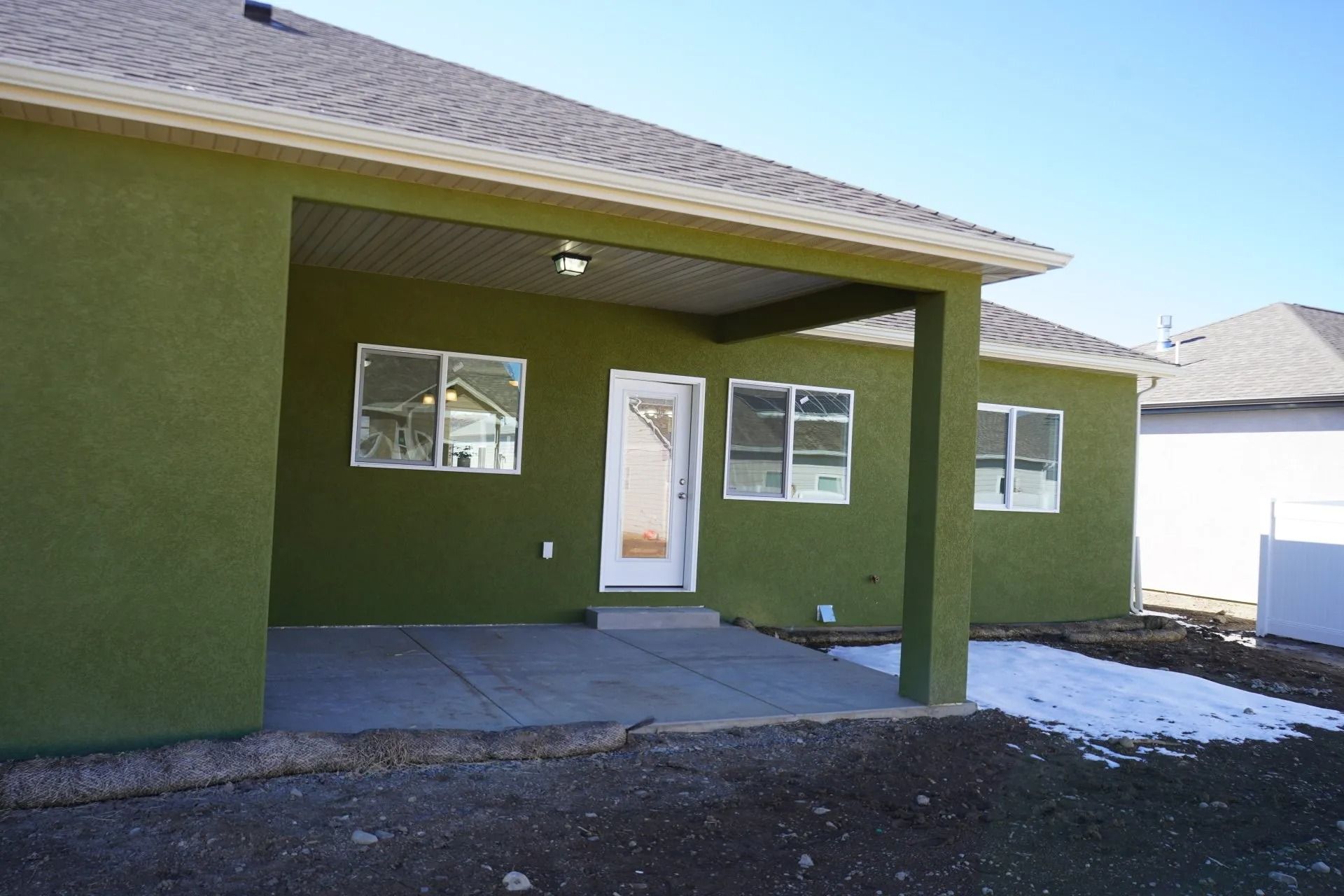 A back view of a green house with a covered concrete patio, sliding glass door, and white windows under a blue sky.