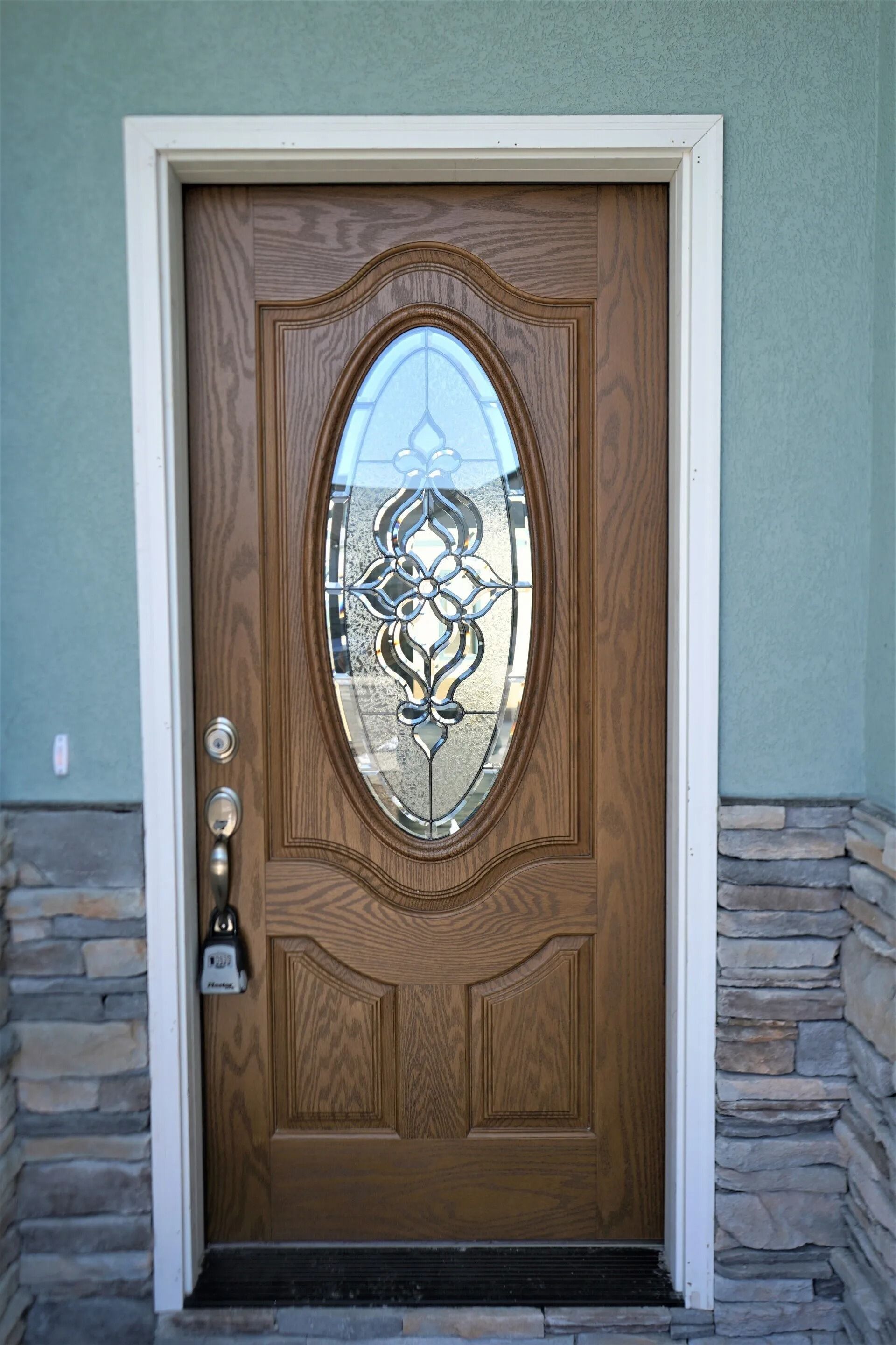 A wooden front door with an oval glass window, set in a light blue wall with stone siding at the base.
