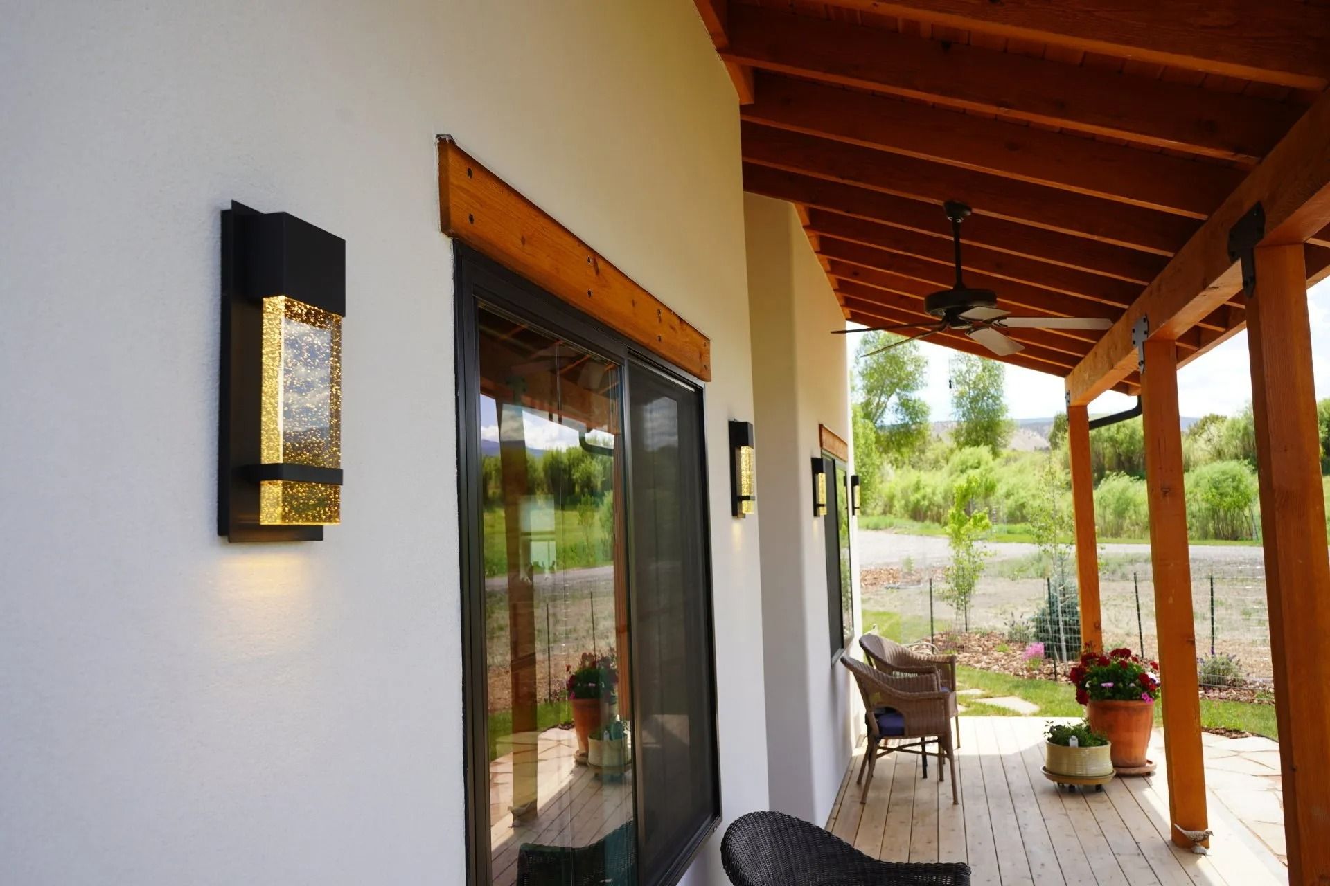 A covered patio with white walls, wooden ceiling beams, outdoor lighting, and wicker chairs overlooking a rural landscape.