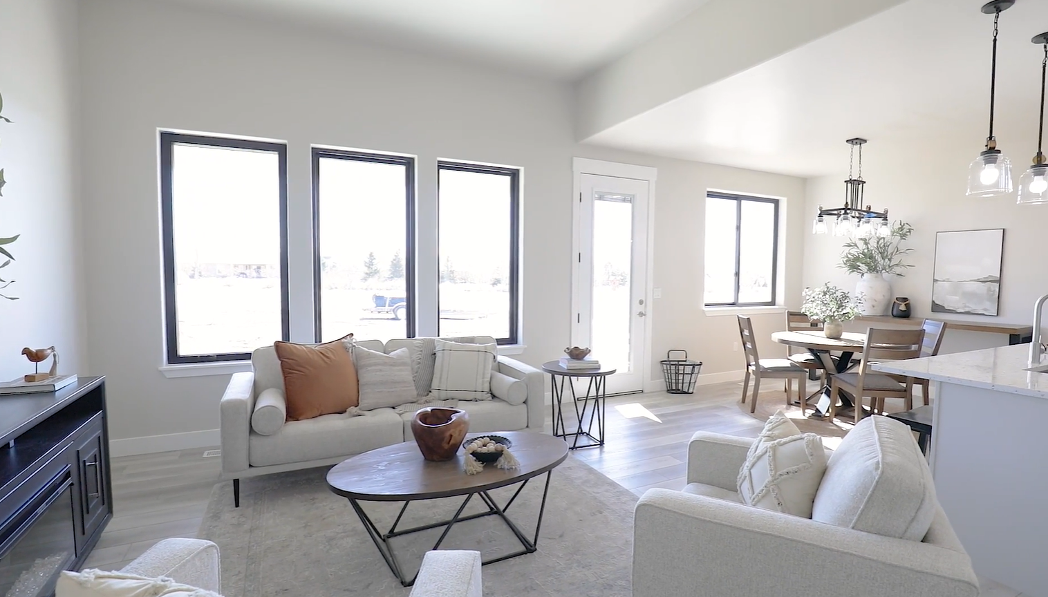 A bright, modern open-concept living and dining area with neutral furniture, a round wood coffee table, and large windows.