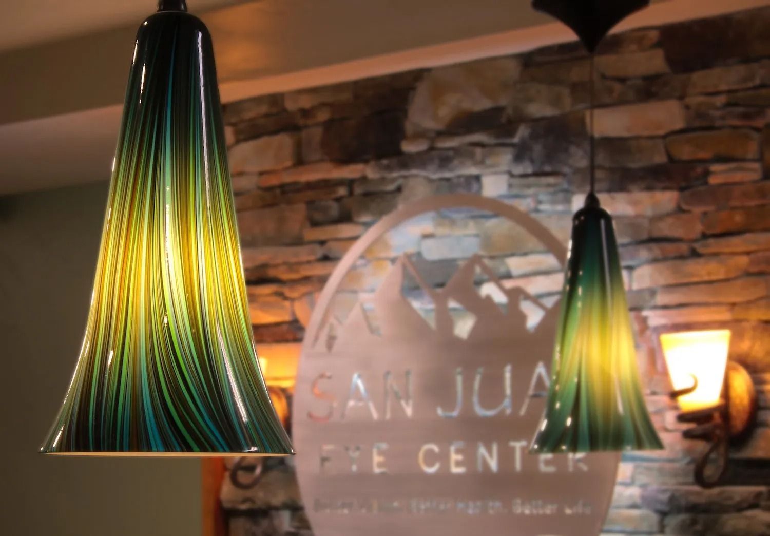 Two hanging glass lamps with green and yellow swirl patterns, in front of a San Juan Eye Center sign on a stone wall.