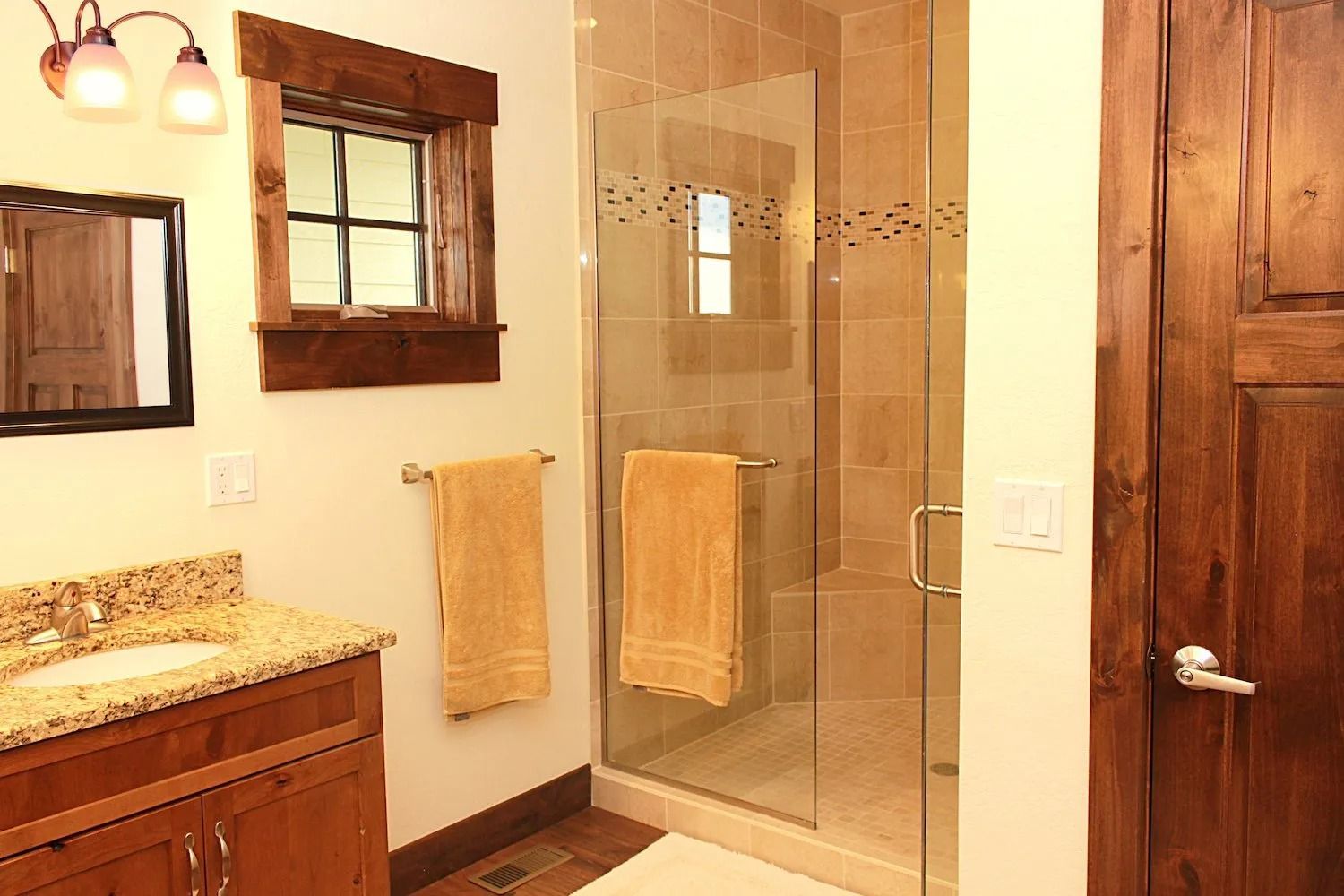 A bathroom featuring a wooden vanity, a granite countertop, a window with dark wood trim, and a glass-enclosed shower.