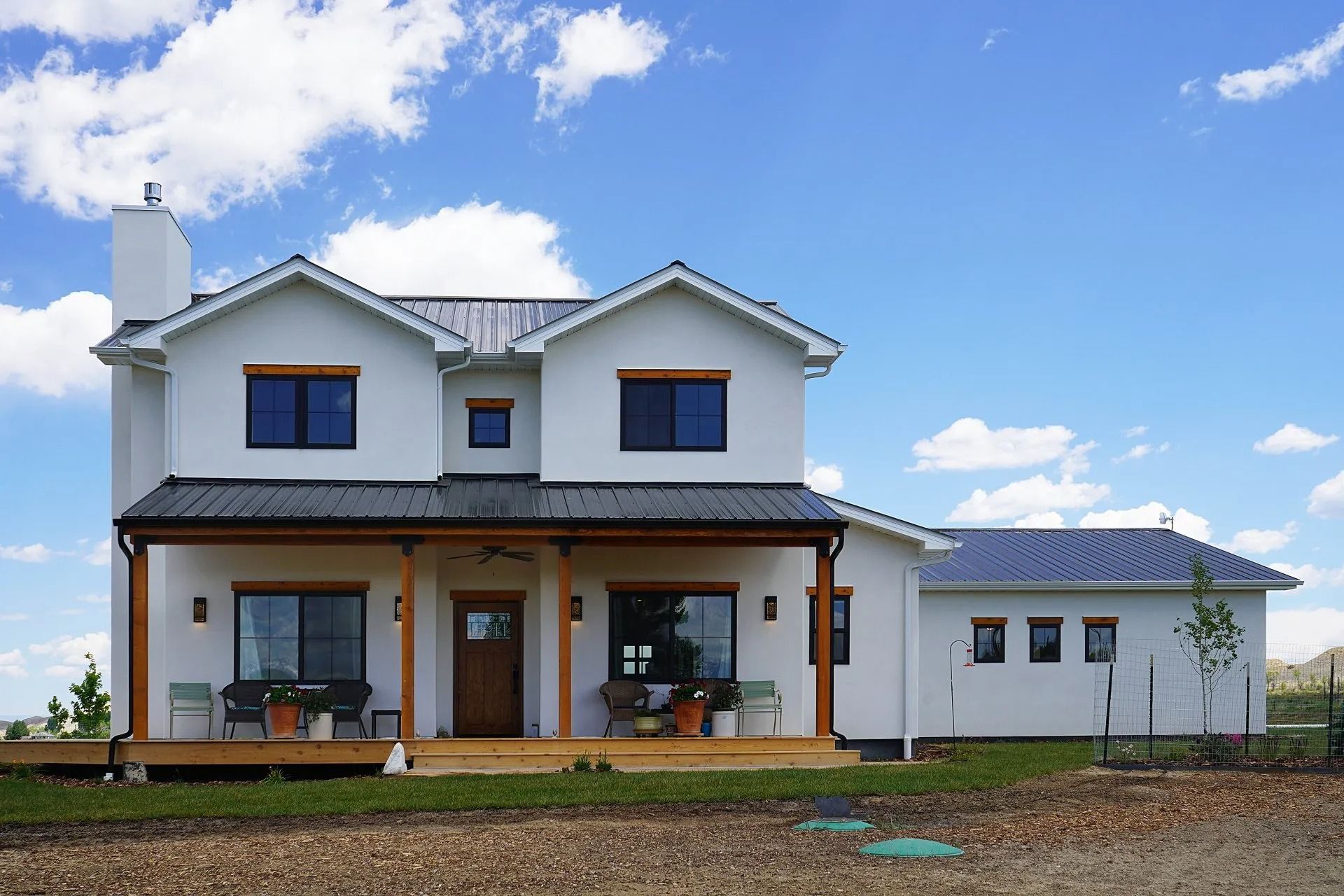 A two-story white farmhouse with a metal roof and large covered front porch under a blue sky with scattered clouds.