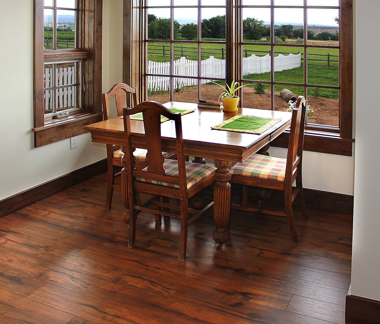 A wooden dining table with four chairs and green placemats, situated in a room with large windows overlooking a yard.