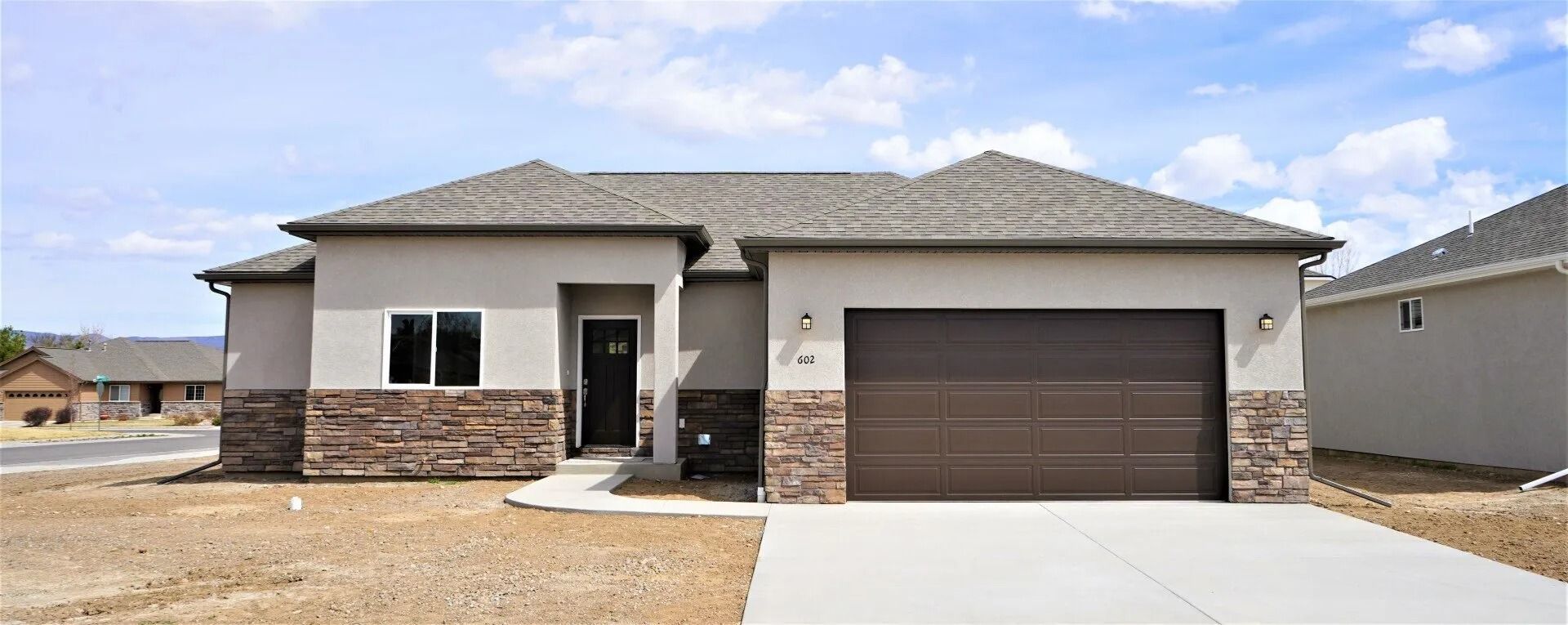 A single-story suburban house with beige stucco, stone veneer accents, a dark garage door, and an unfinished yard.