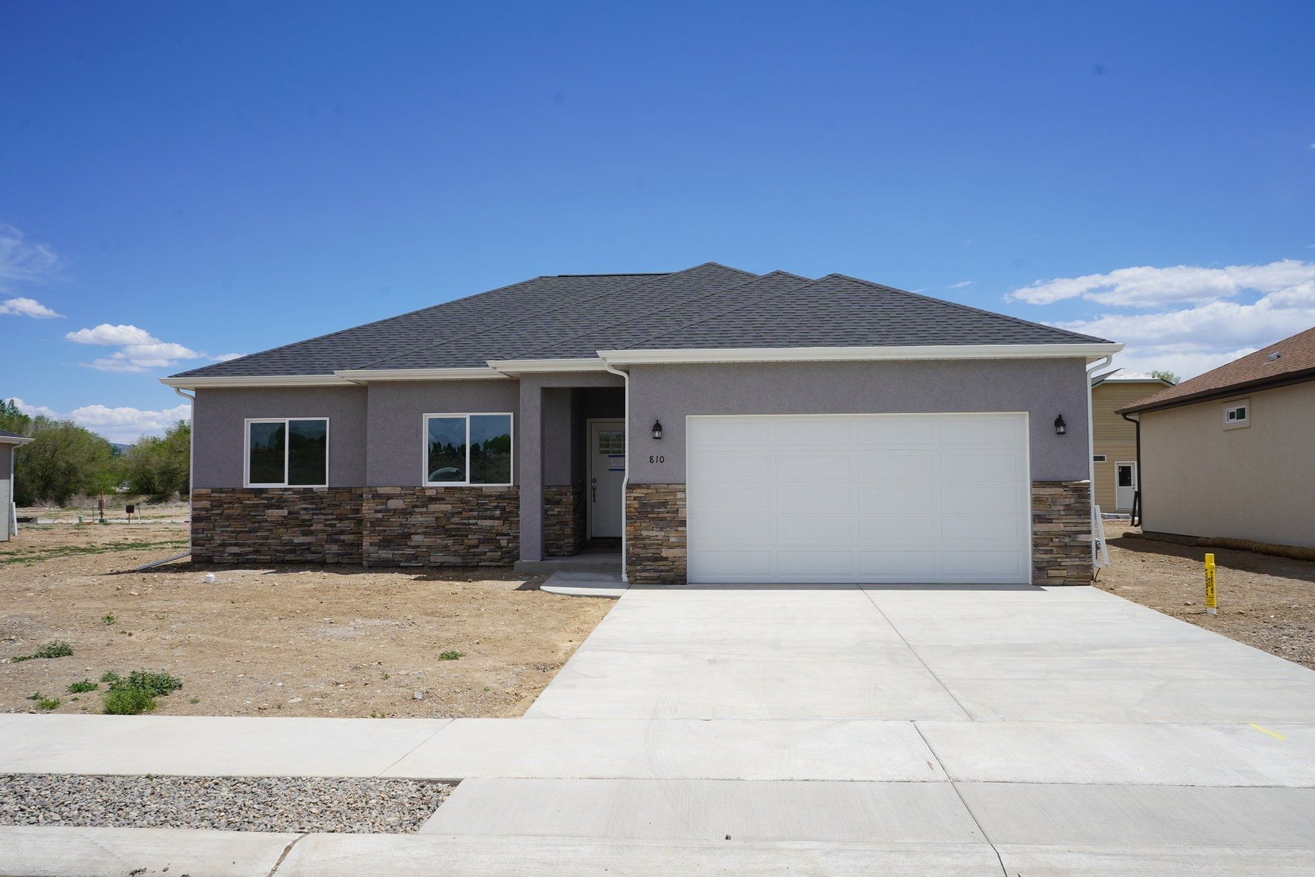 A single-story gray house with stone accents, a two-car garage, and a concrete driveway under a clear blue sky.
