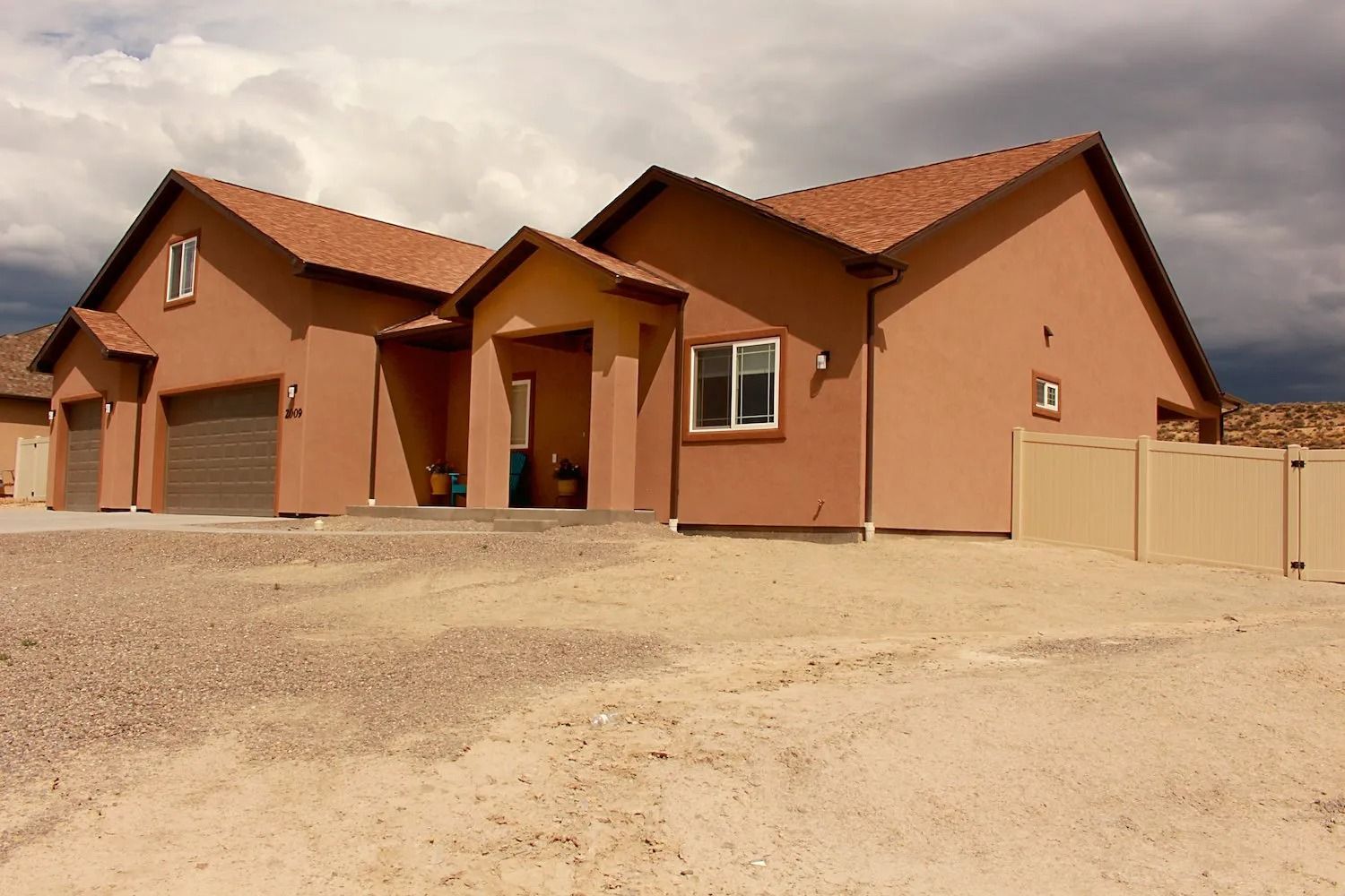 A tan, single-story house with a brown tile roof and gravel front yard under a cloudy sky.