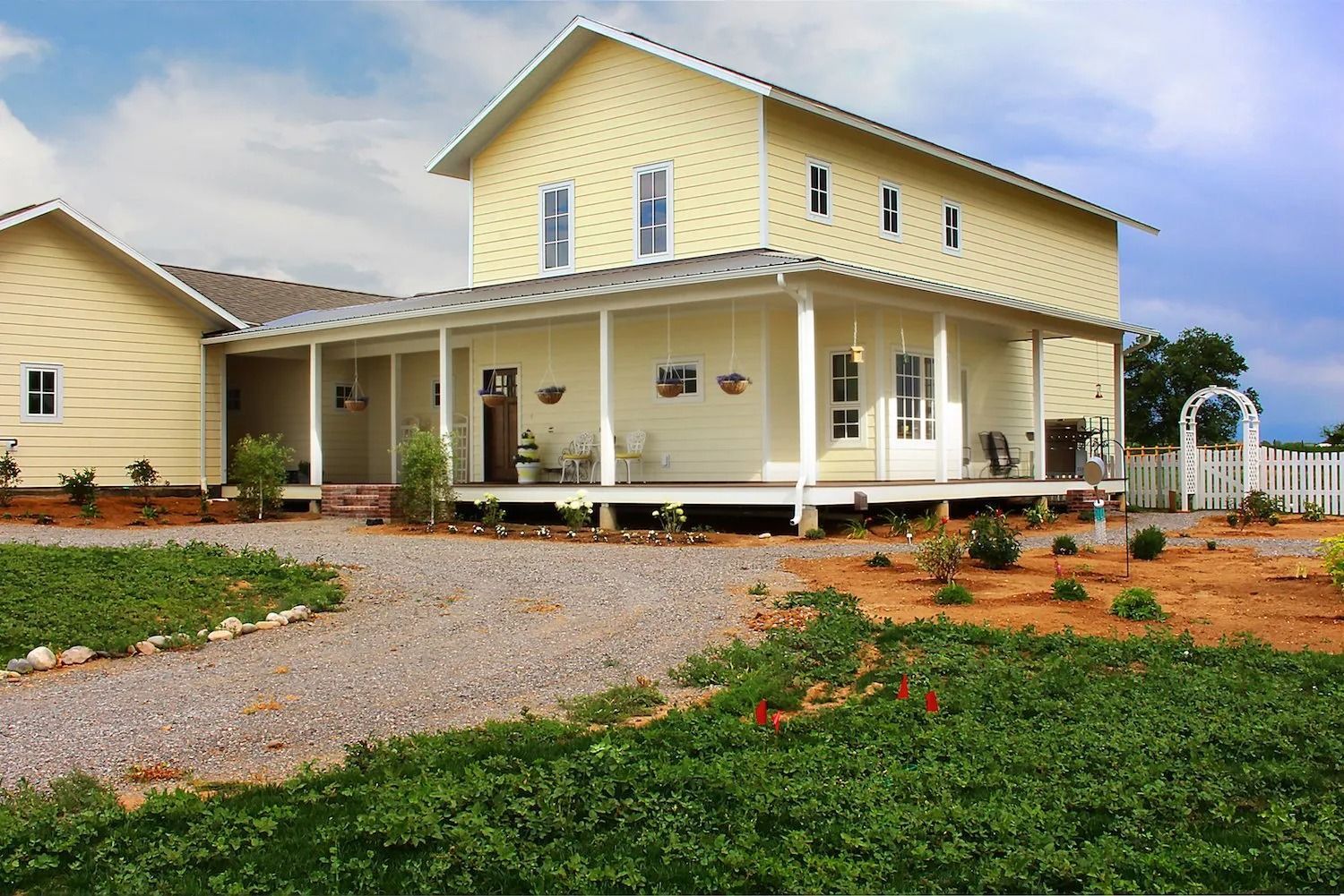 A two-story light yellow house with a sprawling covered porch sits behind a gravel driveway under a bright blue sky.