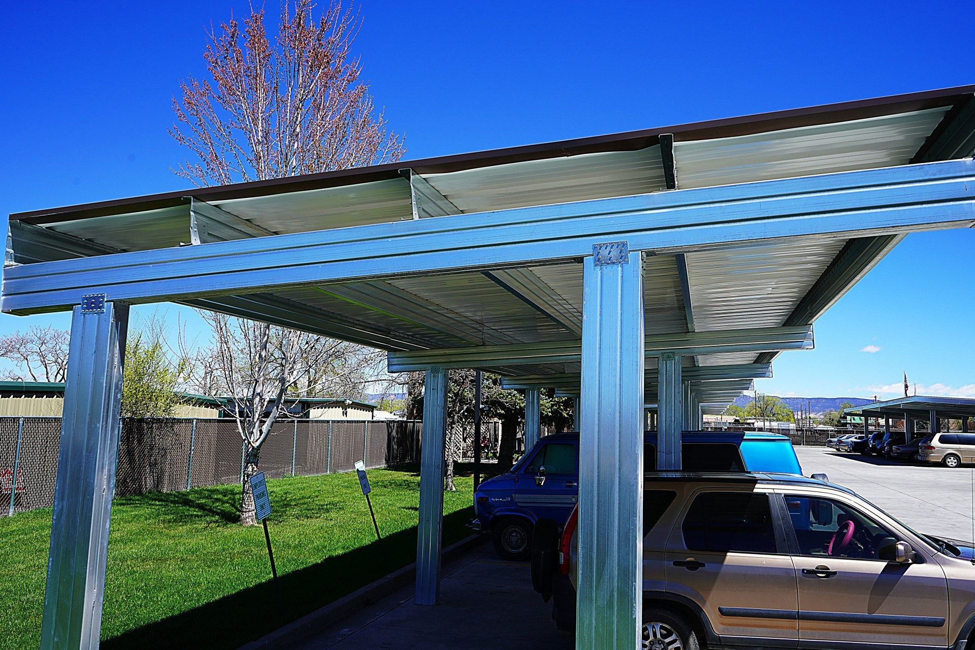 A steel-framed parking canopy structure stands in a parking lot under a clear blue sky on a sunny day.