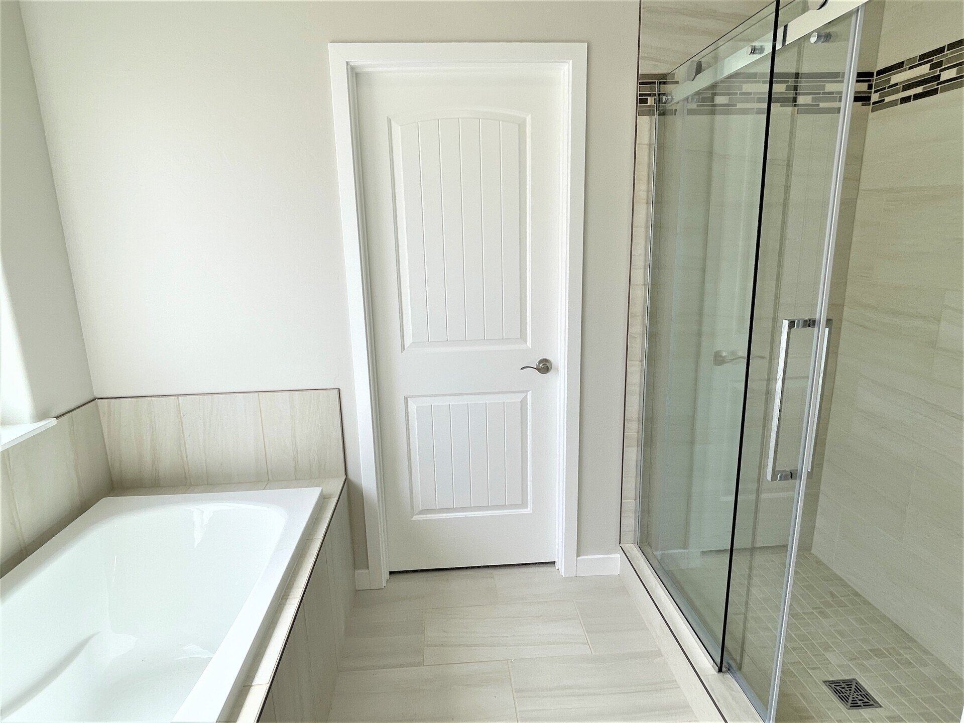 A modern bathroom featuring a white soaking tub on the left, a central white door, and a glass-enclosed shower on the right.