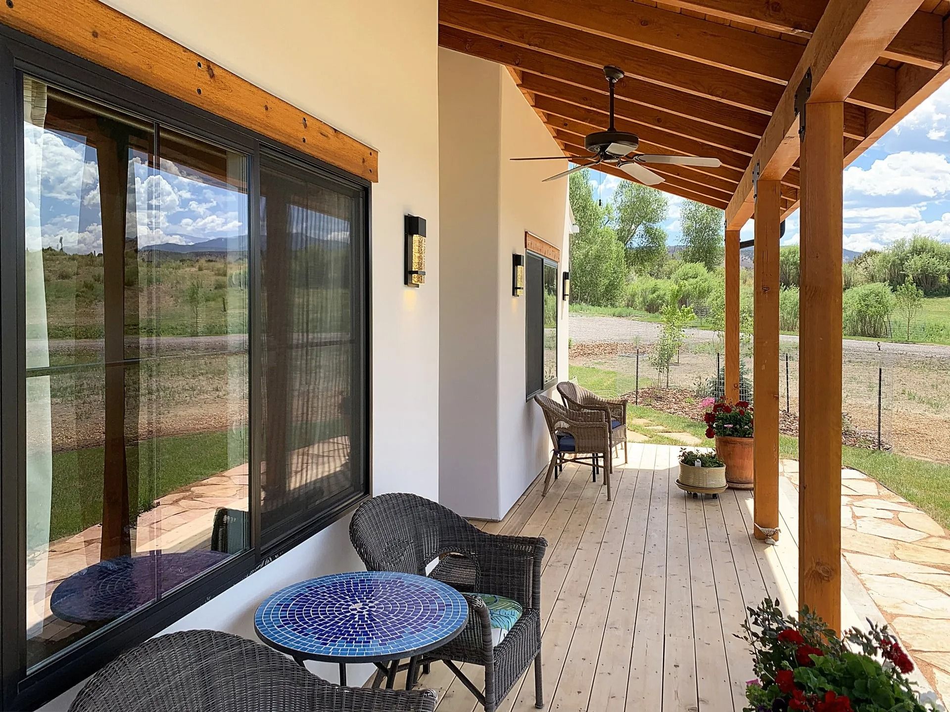 A covered patio with a wooden ceiling, wicker chairs, a small mosaic table, and a view of a rural landscape.