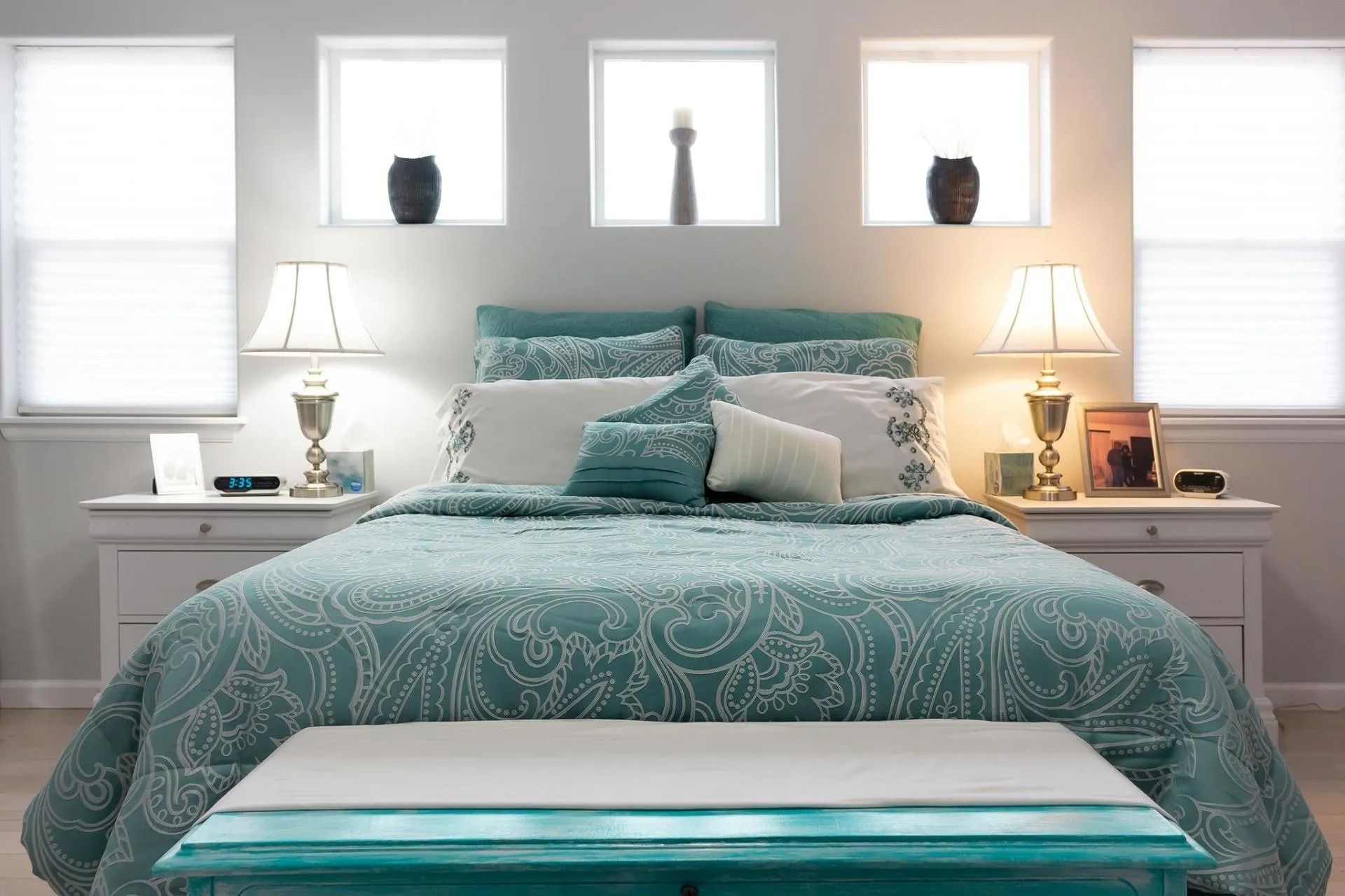 A centered bed with teal paisley bedding, two lamps on white nightstands, and three decorative wall niches above.