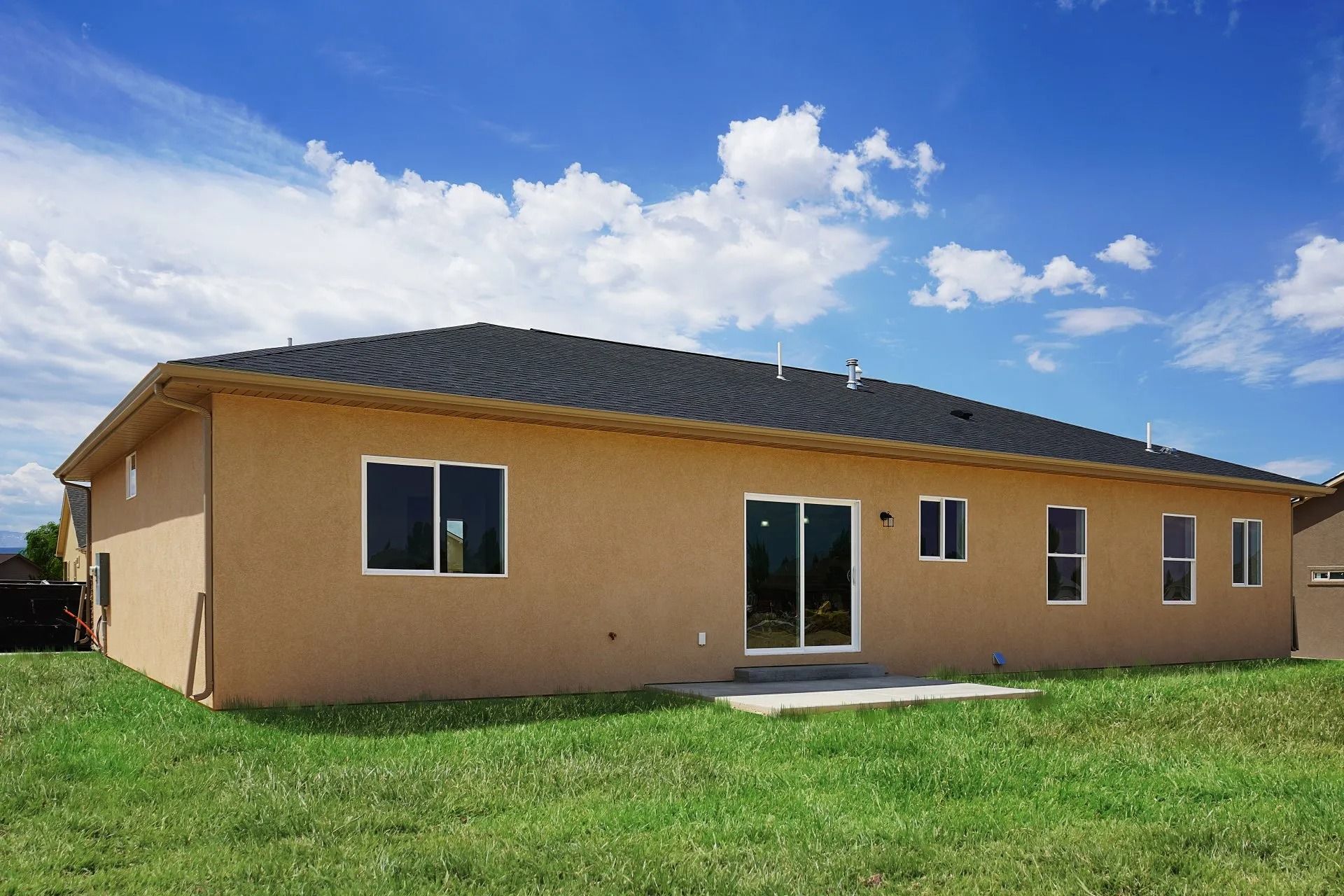 A single-story tan house with a dark roof and multiple windows, viewed from the backyard under a blue, cloudy sky.