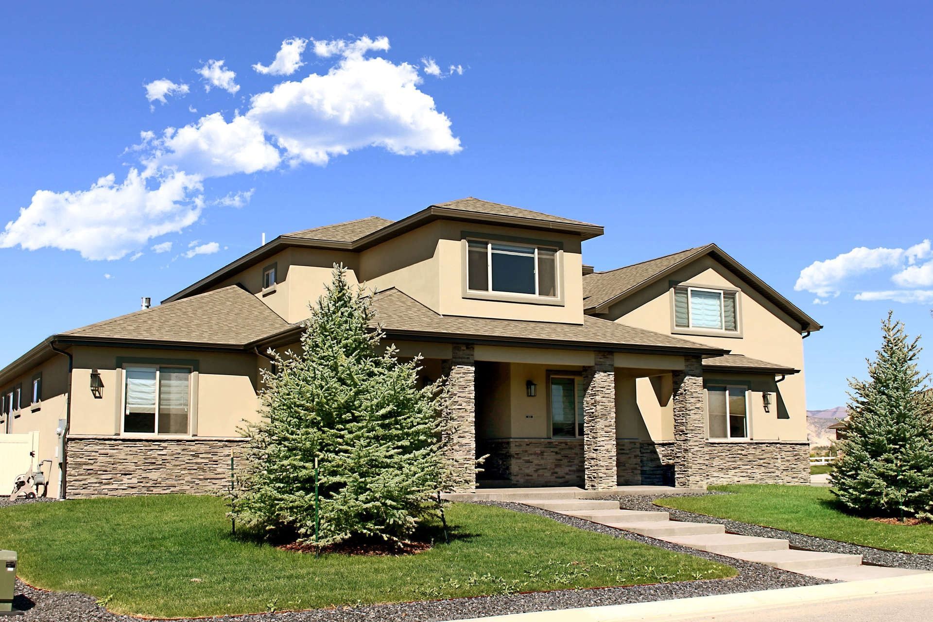 A two-story suburban house with beige stucco walls, stone accents, and two evergreen trees on a grassy lawn under a sky.