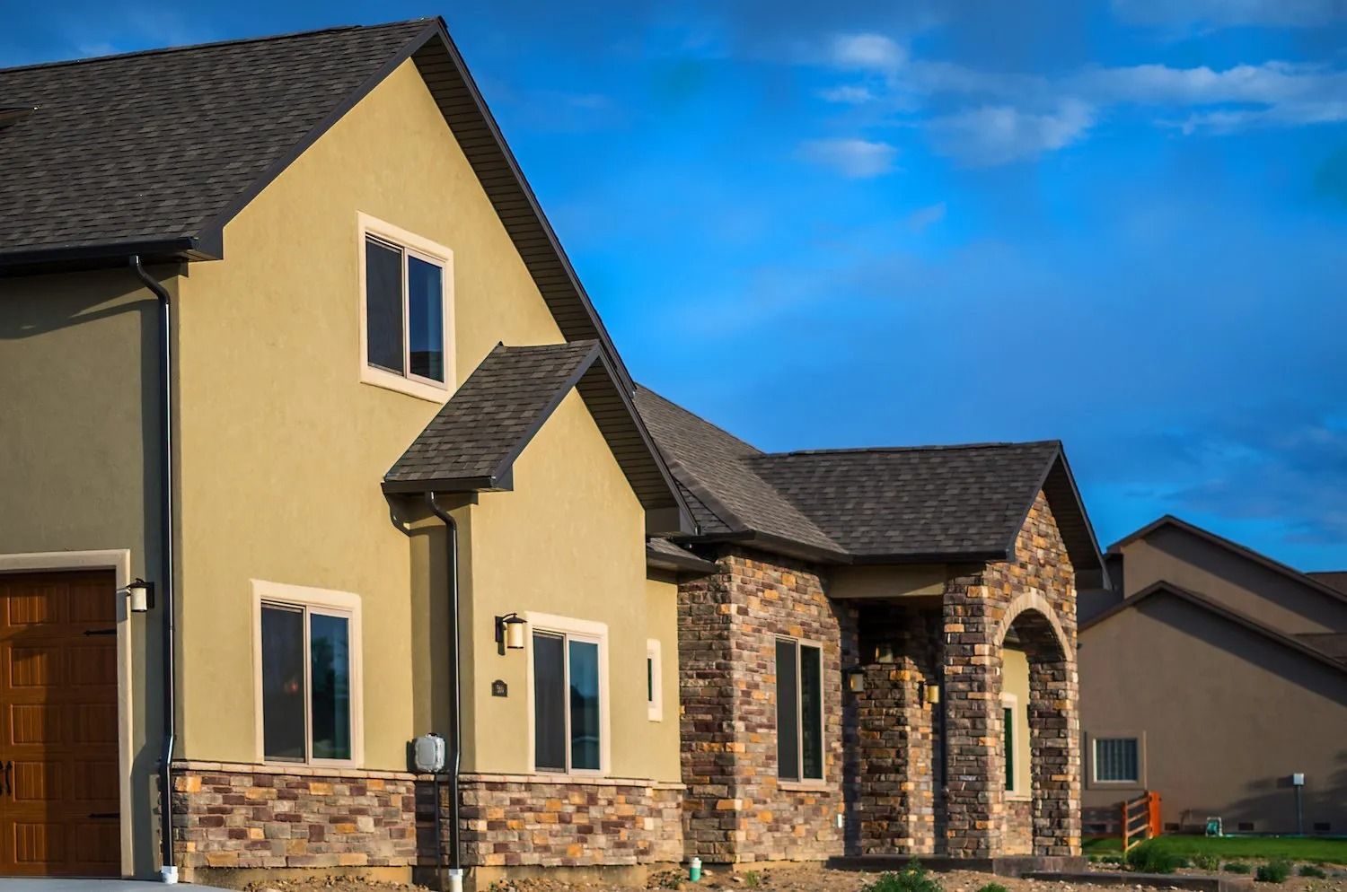 A two-story suburban home with tan stucco walls, stone veneer siding, and a dark shingled roof under a blue sky.
