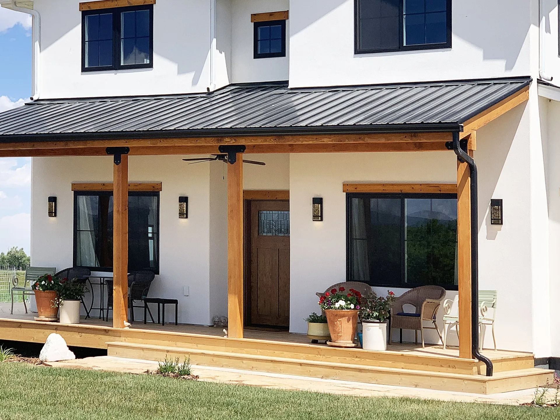 A white house with a dark metal-roofed porch, wooden support beams, a wooden front door, and potted plants on the deck.