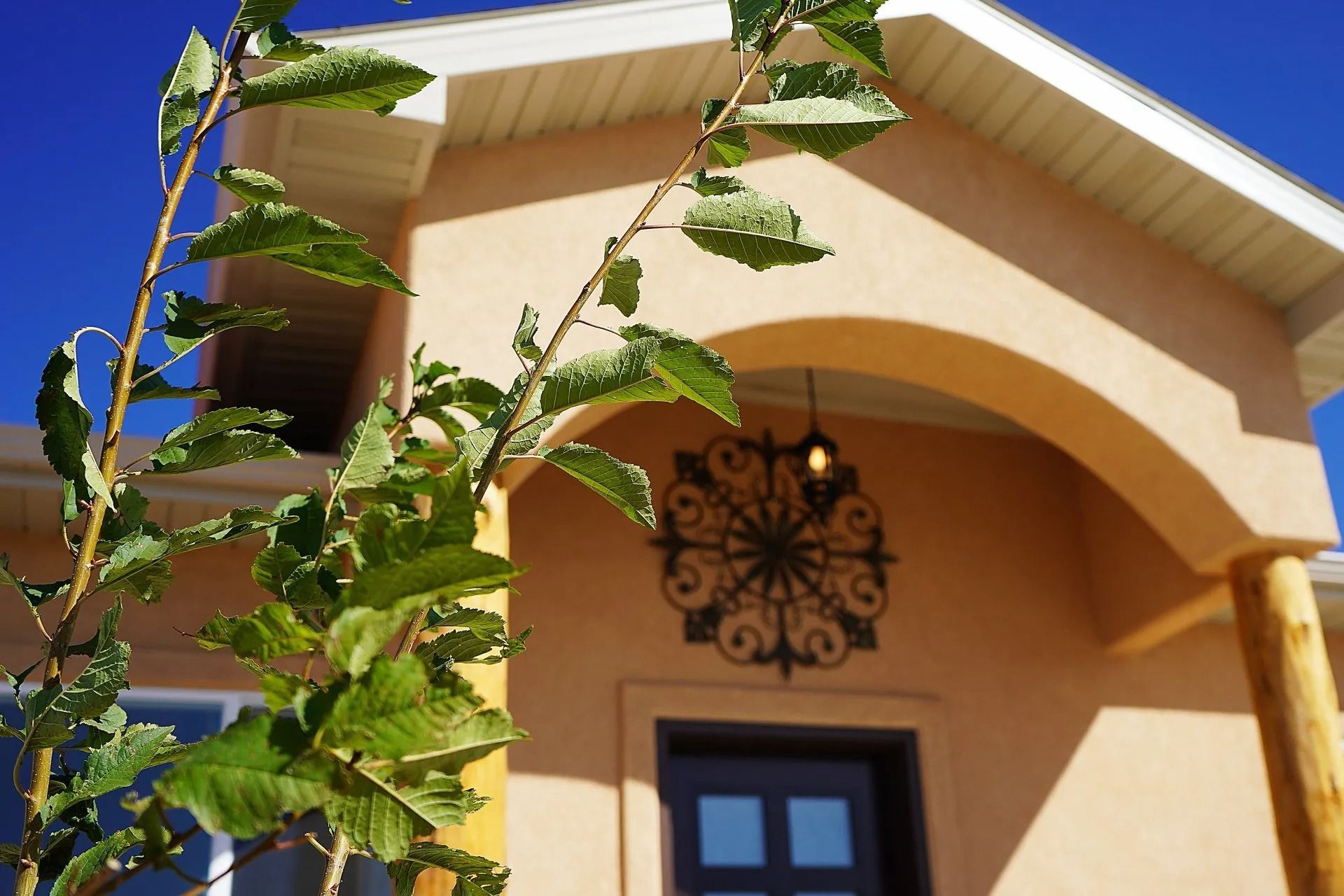 A beige house with a stucco arched entryway, a decorative metal wall hanging, and a green plant in the foreground.