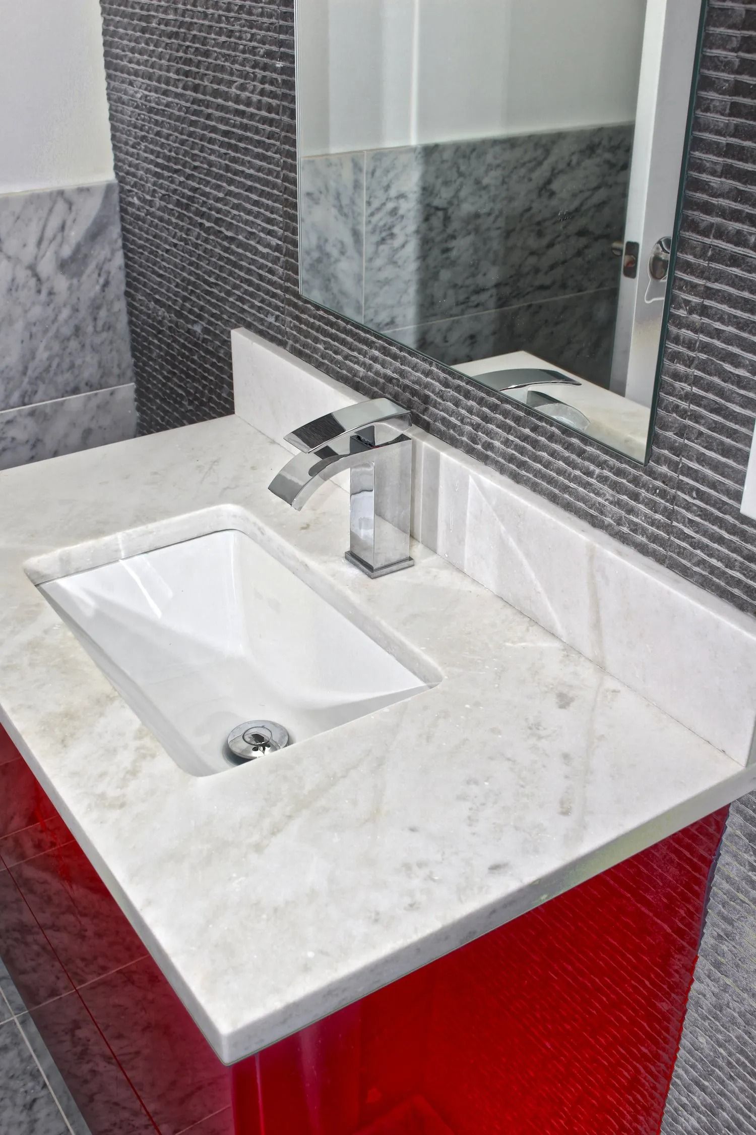 A modern bathroom vanity with a white vessel sink, polished chrome faucet, and a red base against dark textured tiles.