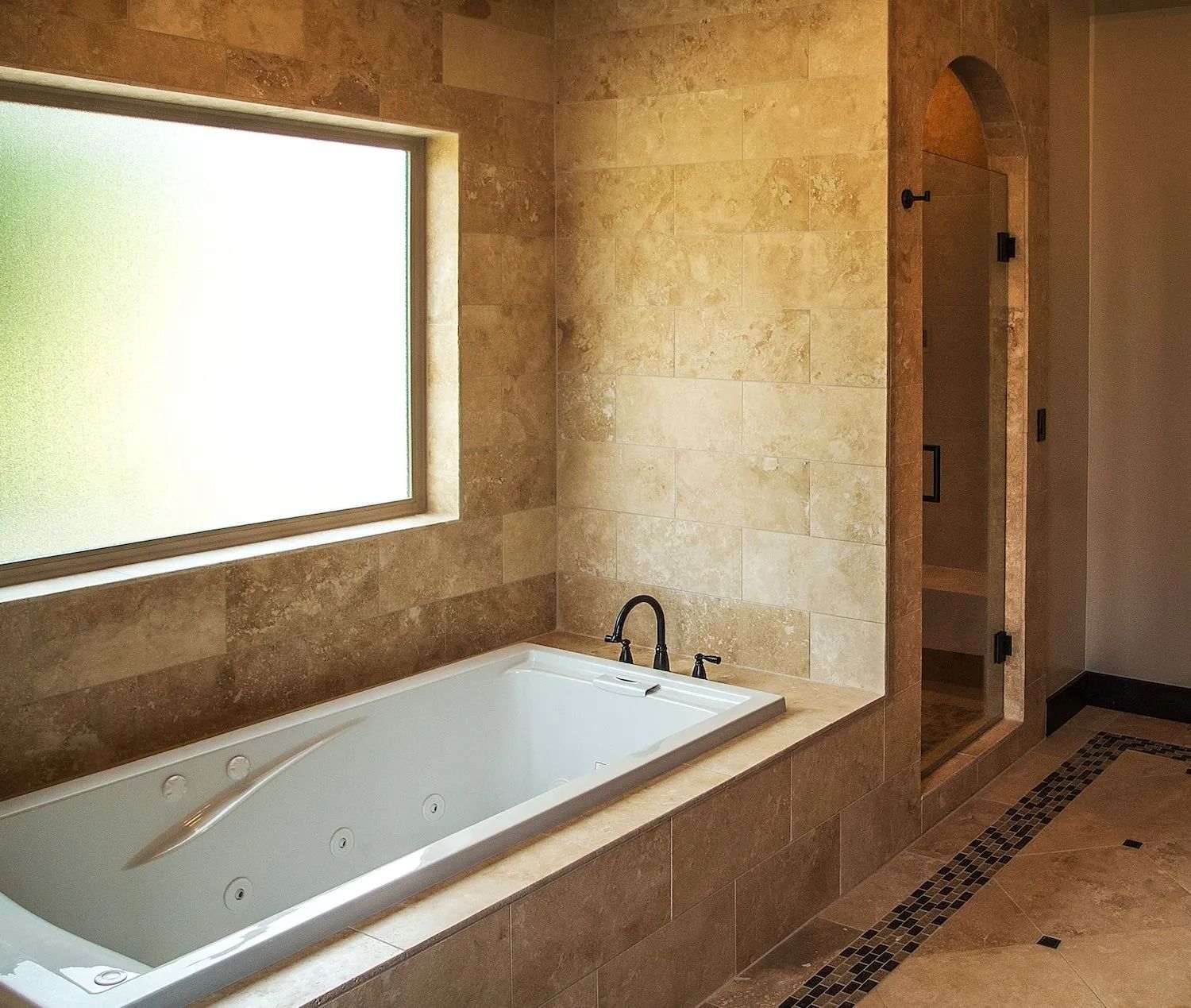 A white soaking tub sits below a window in a bathroom tiled with light brown stone, next to a glass-doored shower.