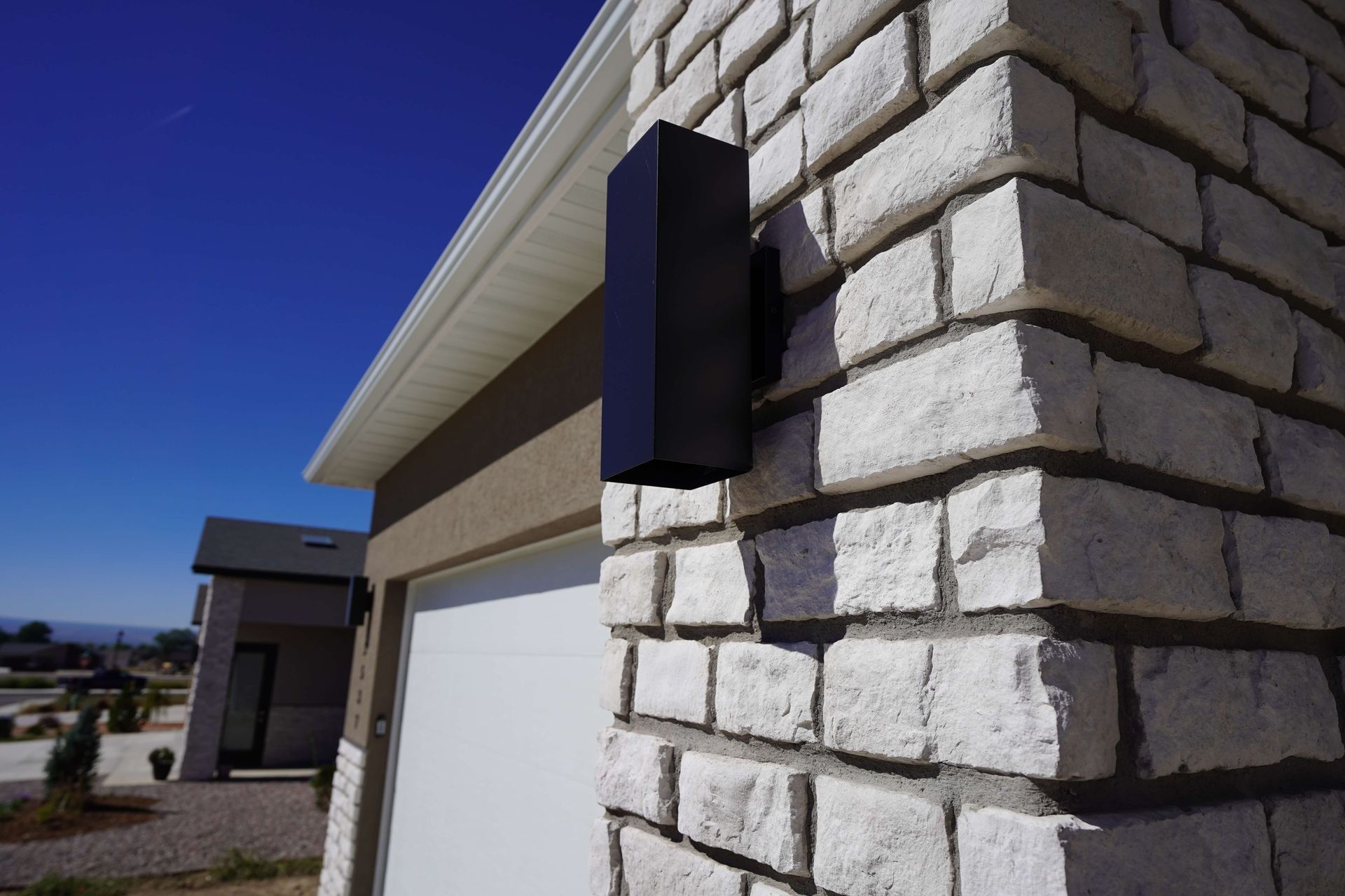 A modern rectangular black light fixture is mounted on a white stone brick wall corner next to a garage under a blue sky.
