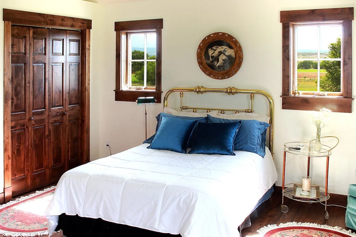 A bedroom with a bed featuring a gold frame, blue pillows, and white bedding, flanked by two windows and a dark wood door.
