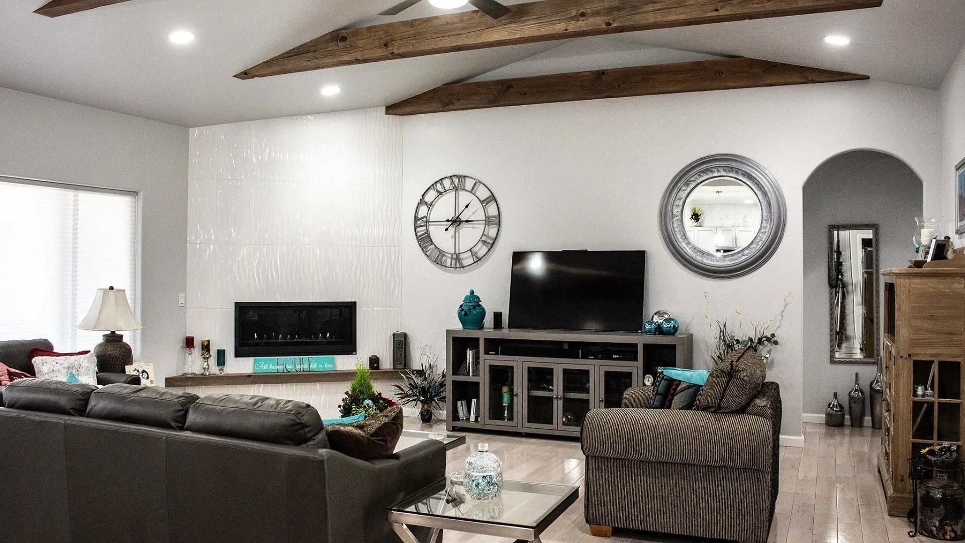 A living room with wooden ceiling beams, a fireplace, a TV, a large wall clock, and a circular mirror above furniture.