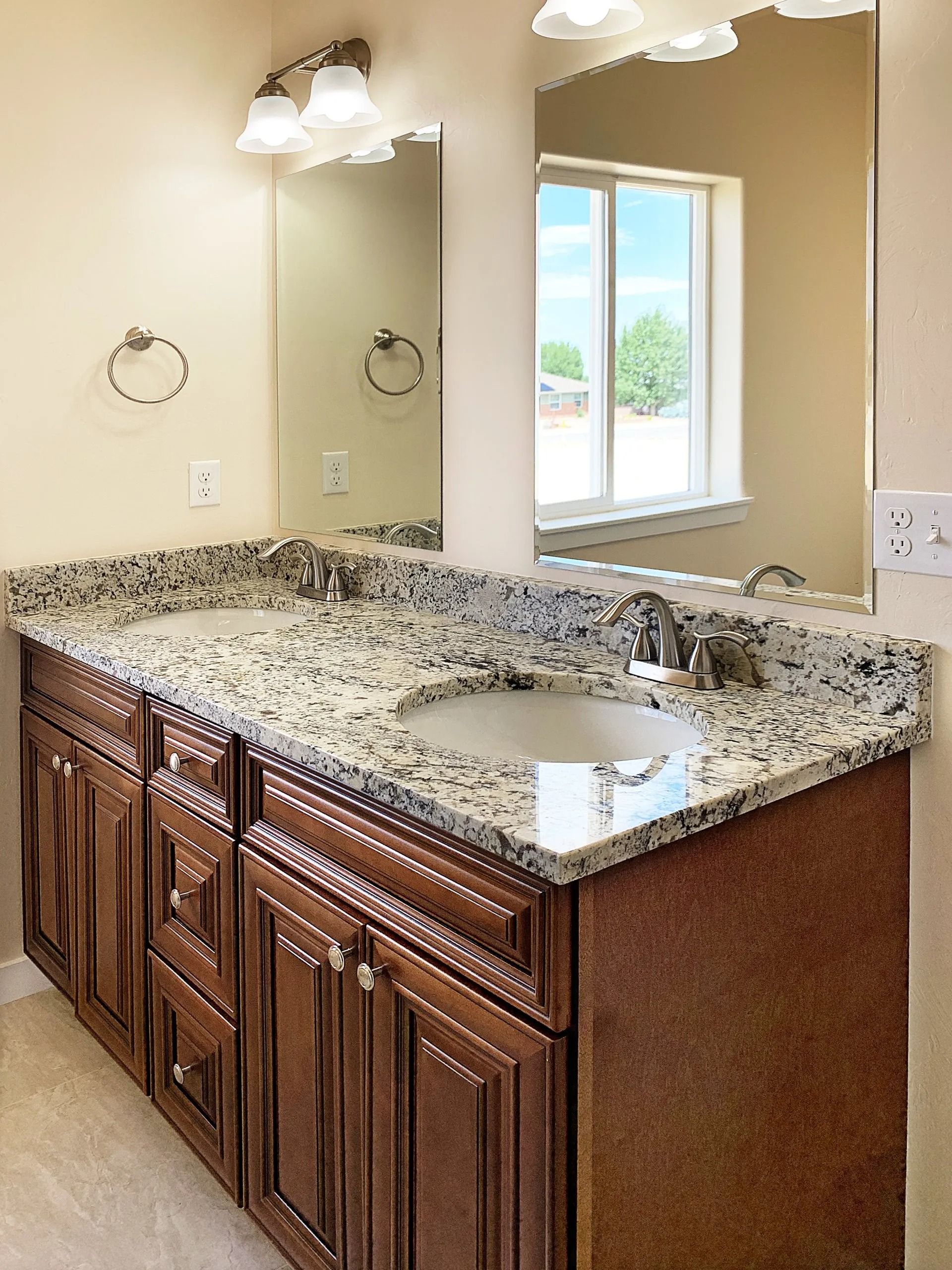 A double-sink bathroom vanity with dark wood cabinets, a speckled granite countertop, and mirrors in a bright room.
