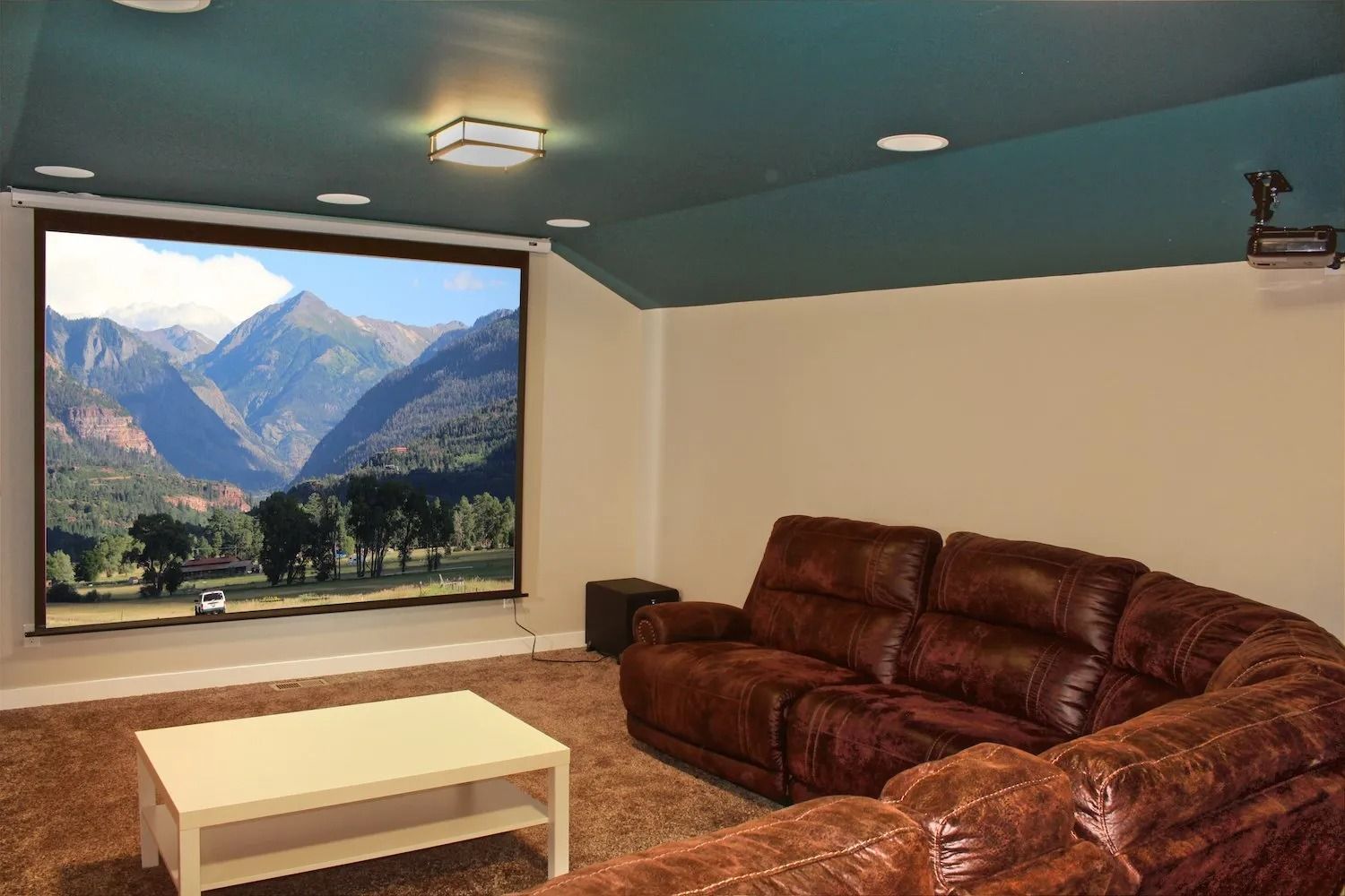 A home theater with a brown leather sectional, a white coffee table, and a large projection screen displaying mountains.