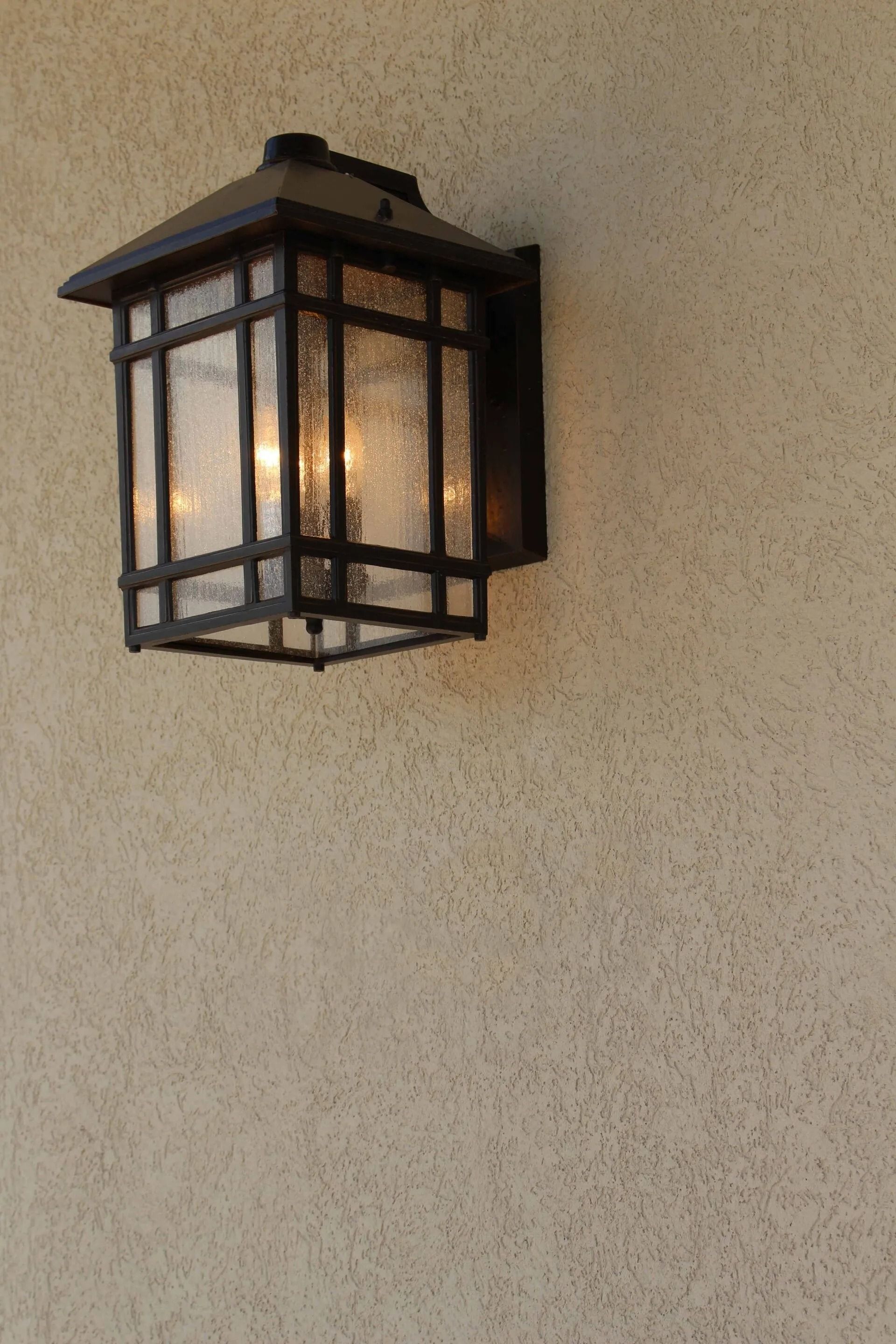 A dark-framed, boxy outdoor lantern with seeded glass panels is mounted on a textured, tan stucco wall.