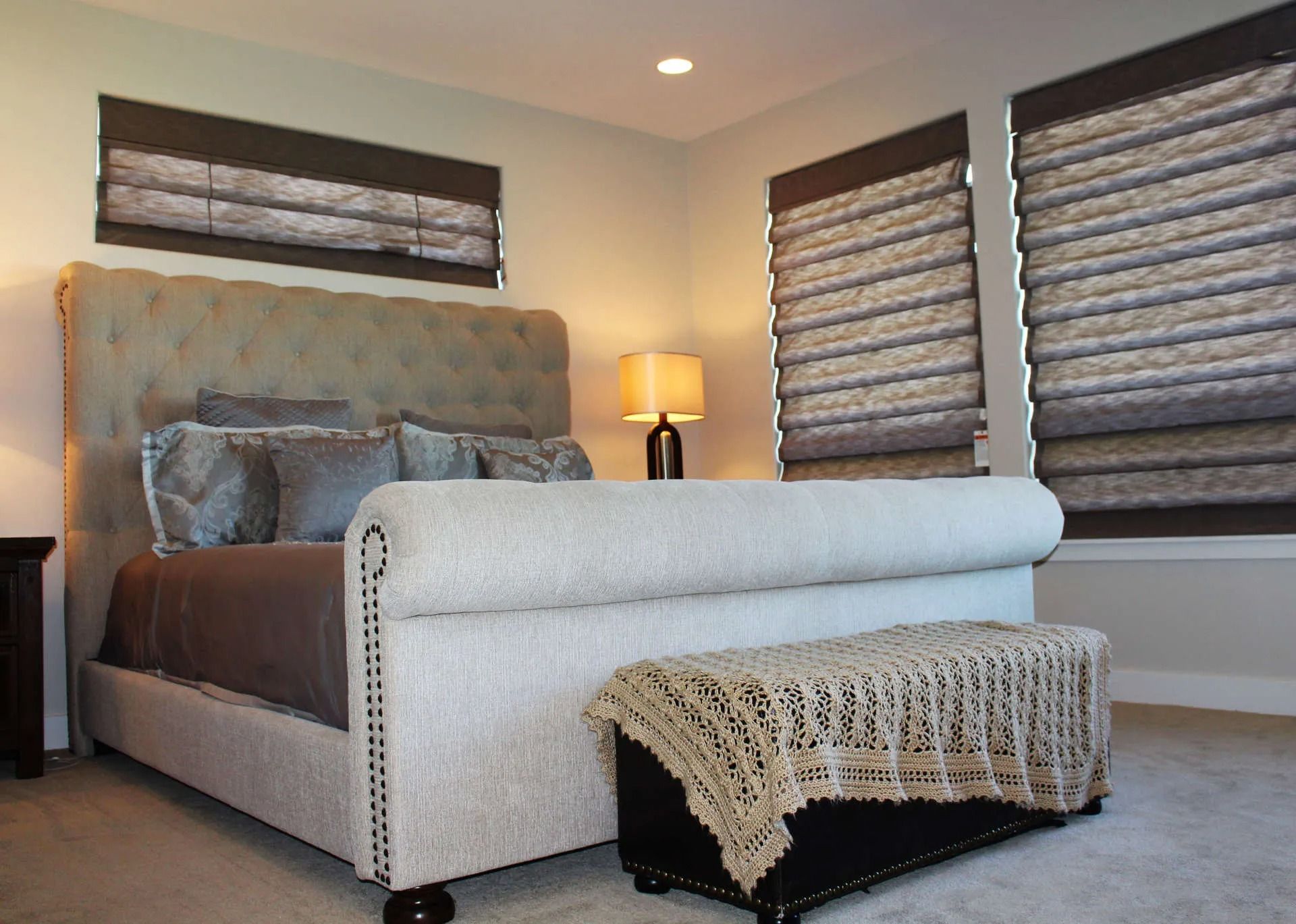 A bedroom with a tufted headboard bed, dark brown bedding, a storage bench, and windows with patterned fabric shades.
