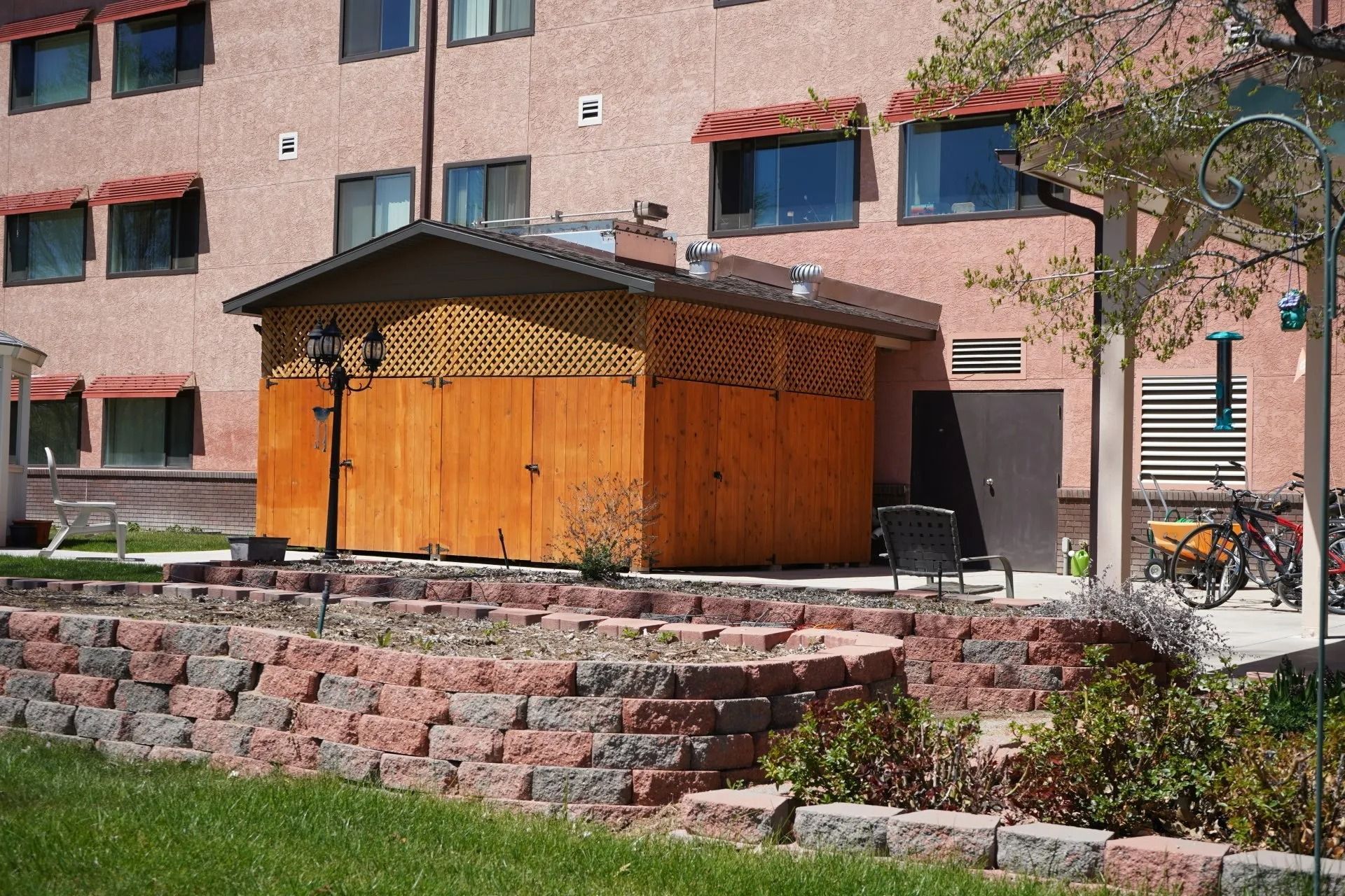 A tan building with a wooden shed structure featuring a lattice top and a low stone retaining wall in front.